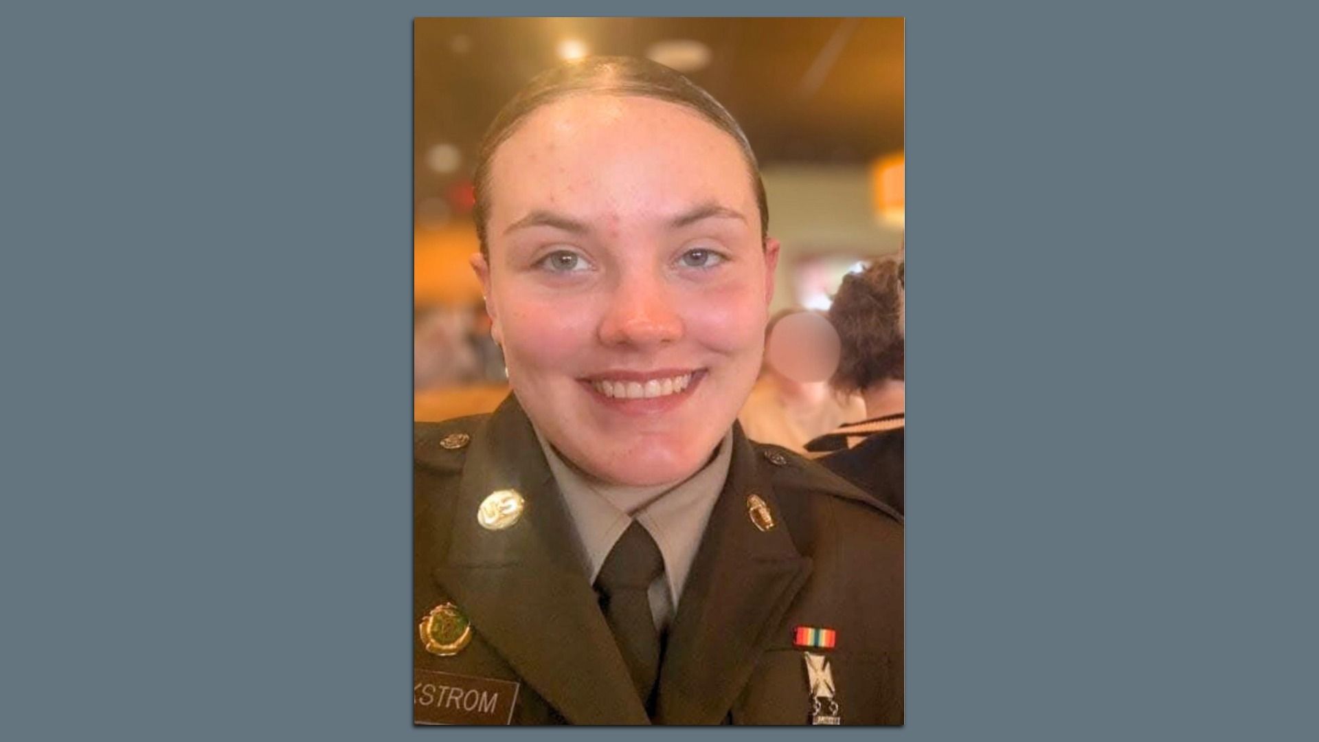 A smiling, dark-haired, fair-skinned West Virginia National Guard member Spc. Sarah Beckstrom, wearing a dark green military uniform with her name tag and various pins and ribbons, against a blurred indoor background with warm lighting.