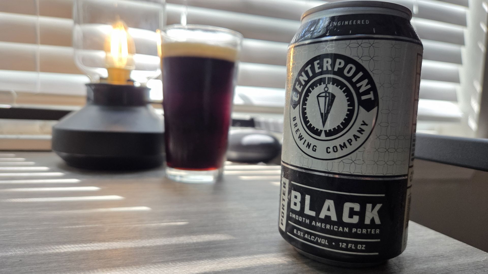 Can of Centerpoint Brewing Company Black Porter beer with a glass of dark beer and a lit lamp on a wooden table near window blinds.