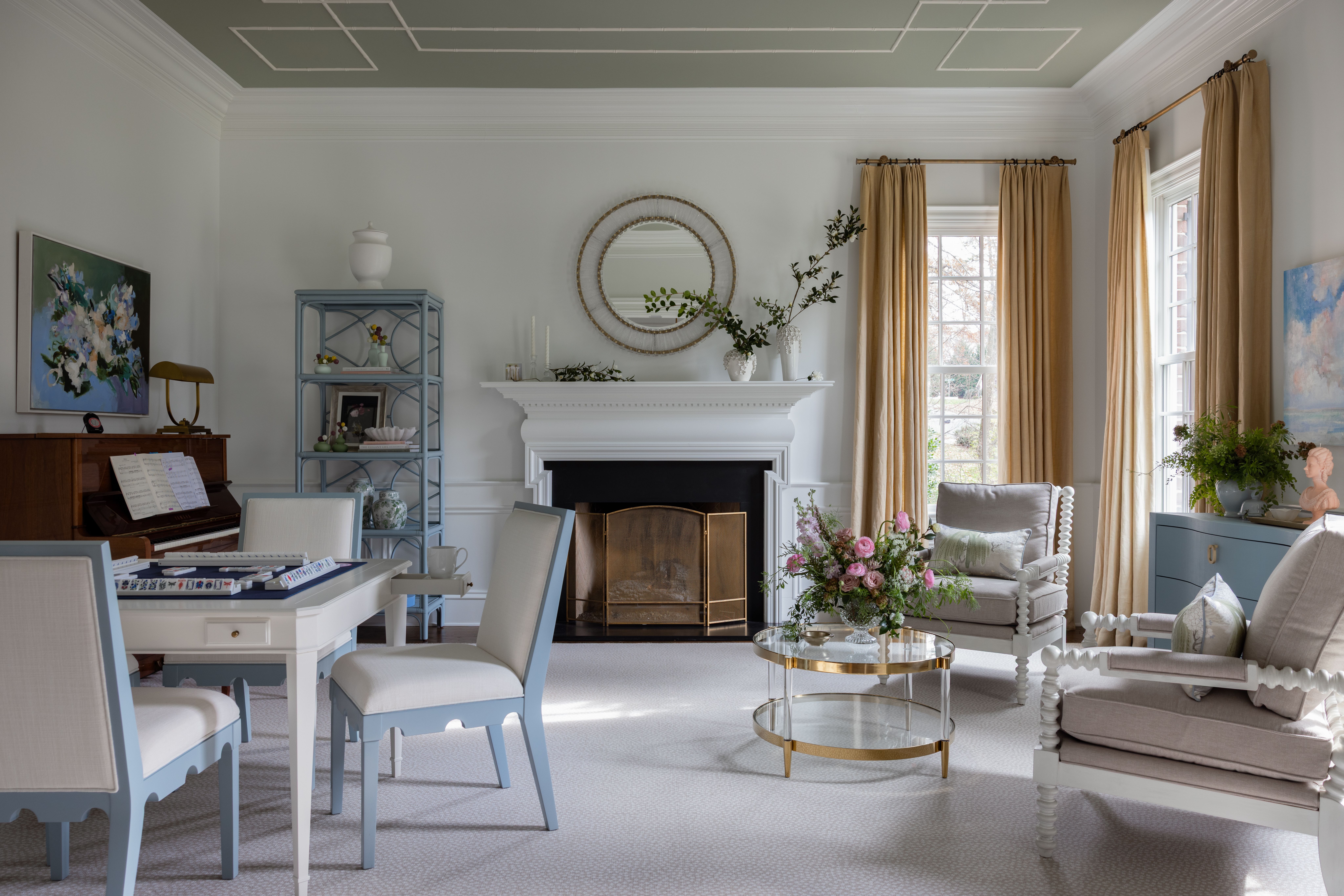 Elegant living room with white walls, beige curtains, two upholstered armchairs, a white and blue table with chairs, a piano, blue shelving, a glass coffee table with flowers, and a fireplace with round mirror.