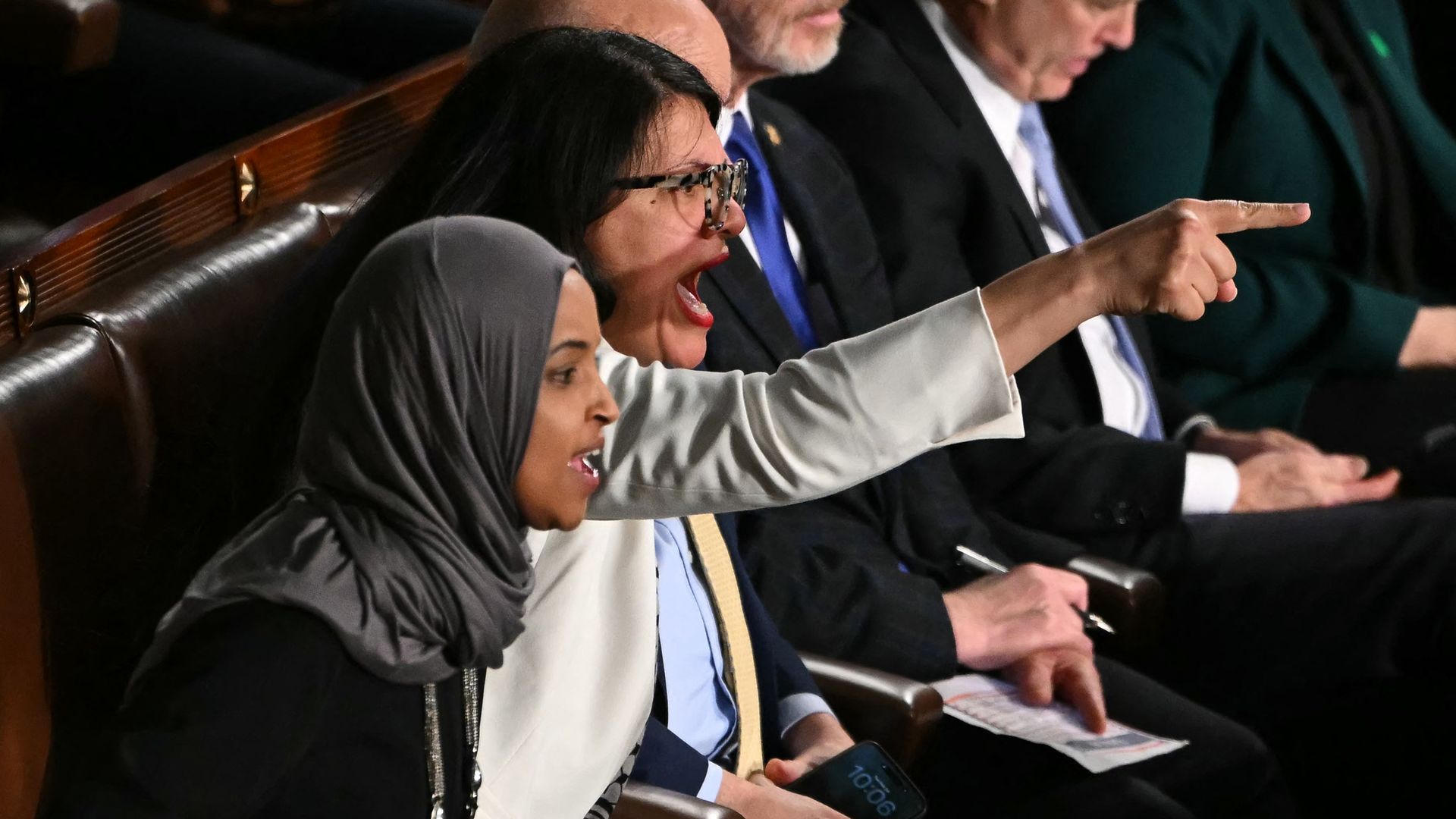 Two women seated, one wearing a gray hijab, the other in a white jacket and glasses, pointing and shouting intensely in a formal setting with people in suits around them.