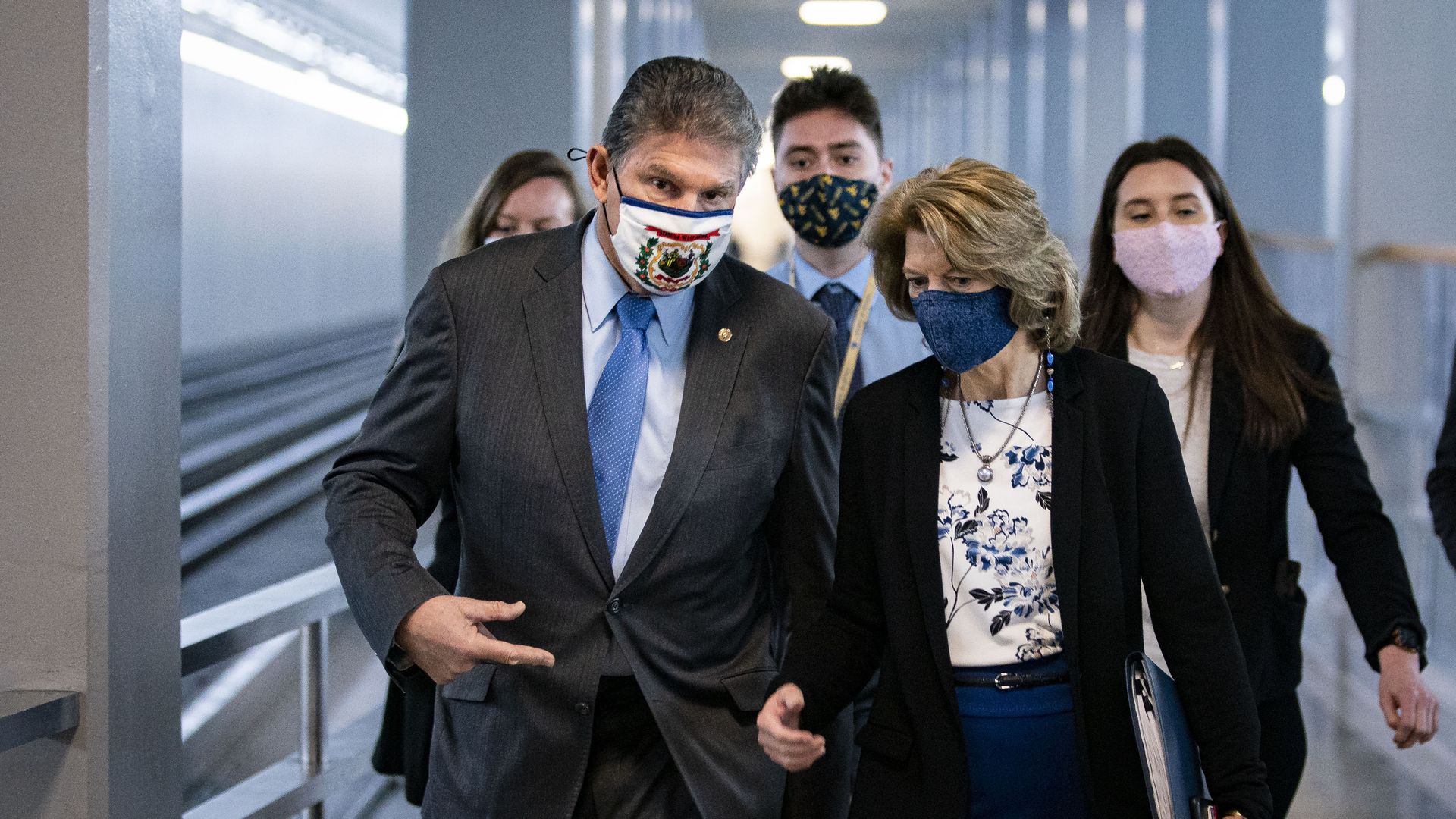 Photo of a masked Joe Manchin and Lisa Murkowski speaking to each other as they walk down a hallway with aides behind them