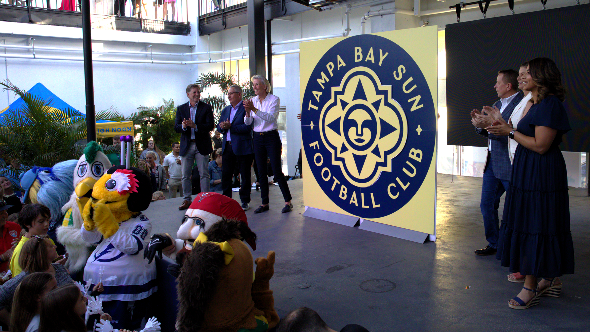 A display board with a blue-and-yellow design of a sun circled by the words "Tampa Bay Sun Football Club." The sign is flaked on either side by three people, all smiling and clapping.