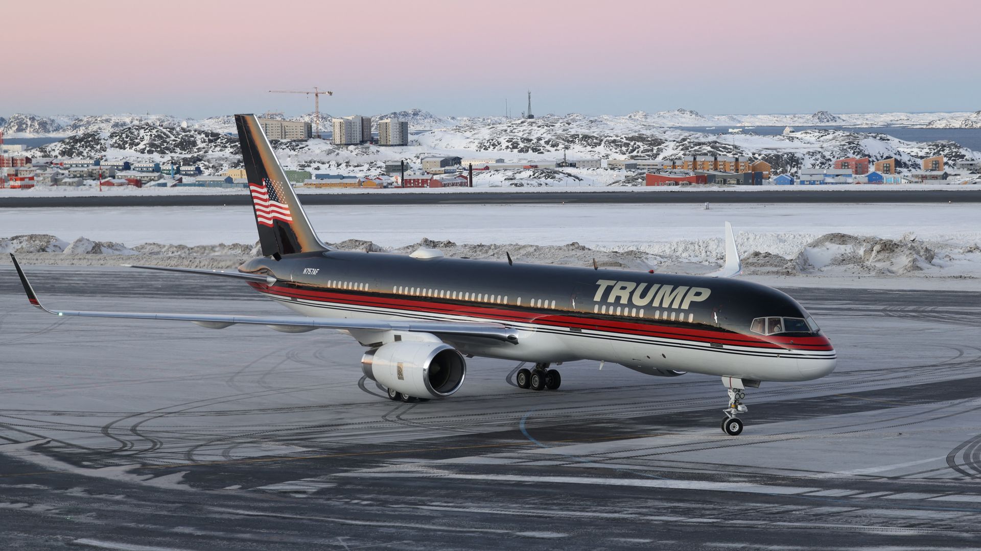 A Trump airplane, dark blue, red and white, stands on an icy runway in Greenland. The sky behind is a mix of pink and blue.