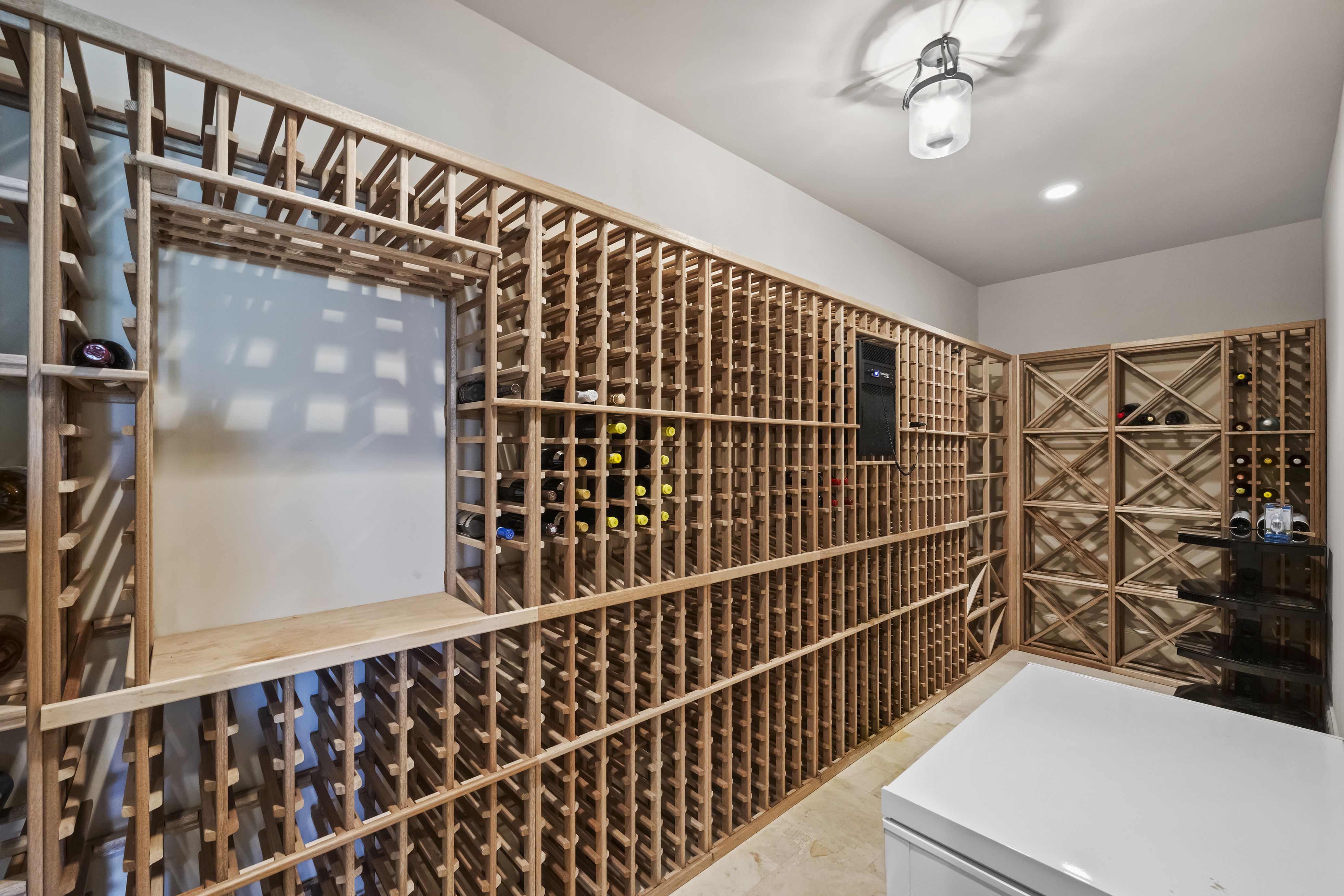 Extensive wooden lattice wine racks line the walls, storing bottles horizontally. White walls, beige floor, and a modern ceiling light. Right side shows glass-front cabinets with more bottles.
