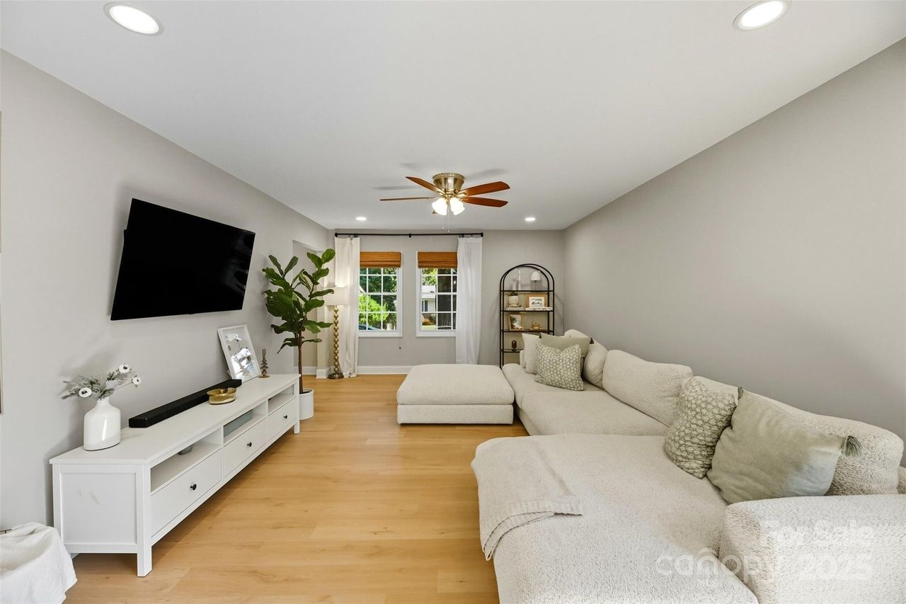 Modern living room with light gray walls, light wood floor, large cream sectional sofa with pillows, white media console, mounted TV, tall plant, and two windows with white curtains.