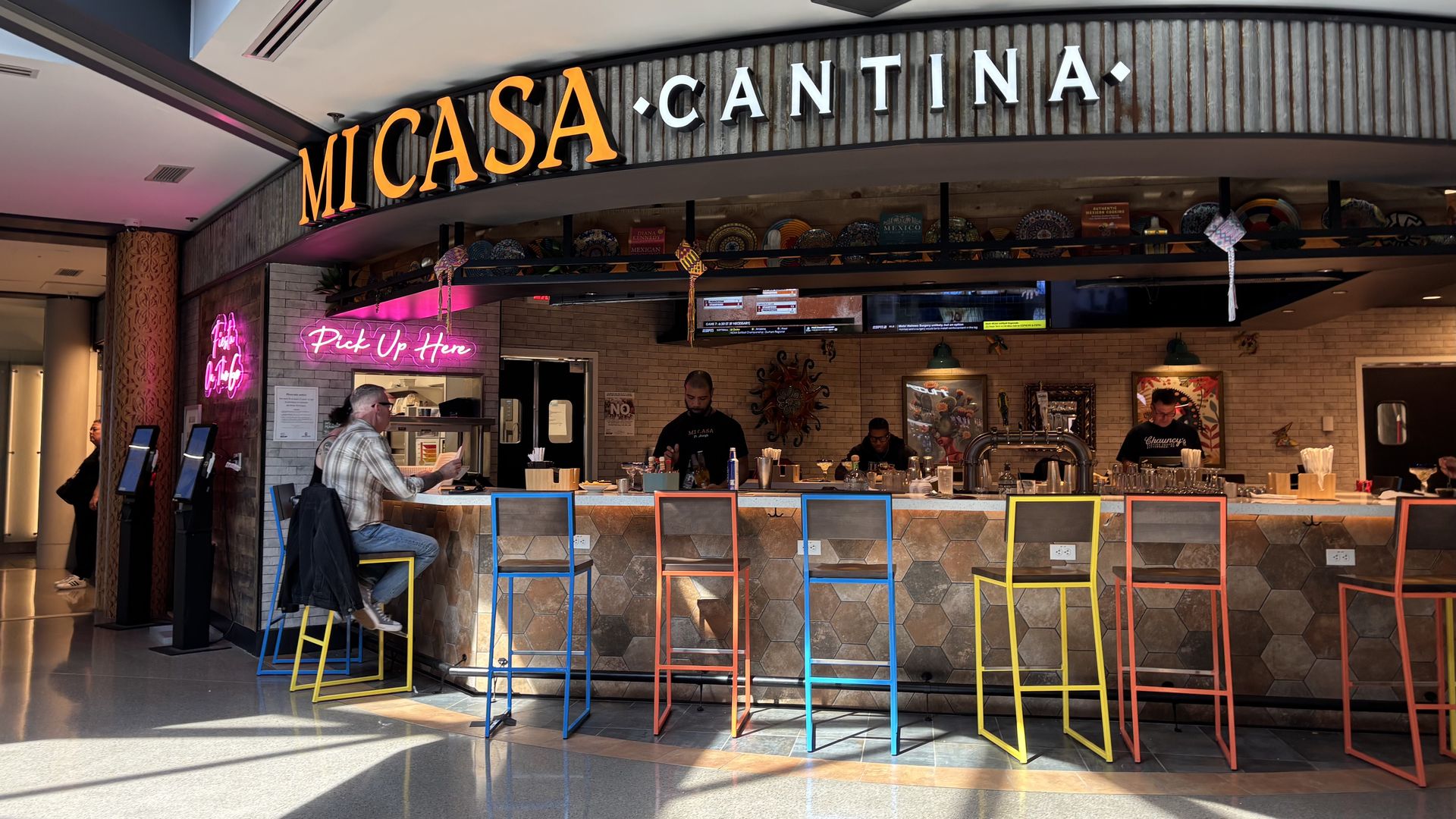 Interior of a casual eatery with a curved counter. A large sign reads "Mi Casa Cantina" on rustic wood. Rainbow stools line the bar, and a pink neon "Pick Up Here" sign glows.