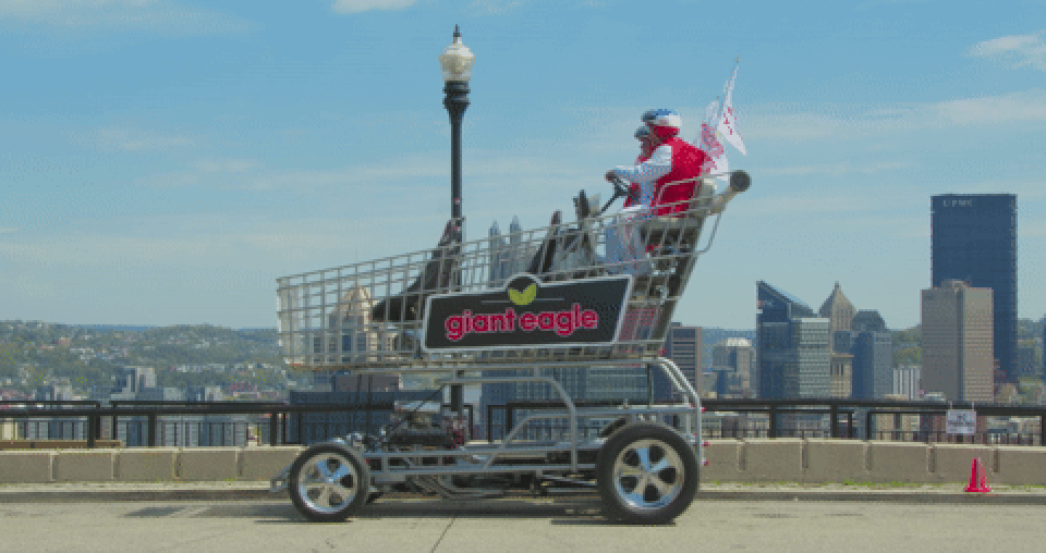 An oversized metal shopping cart on large wheels sits on a waterfront road; two people in red ride inside. City skyline and blue sky backdrop; cart bears the "giant eagle" logo.