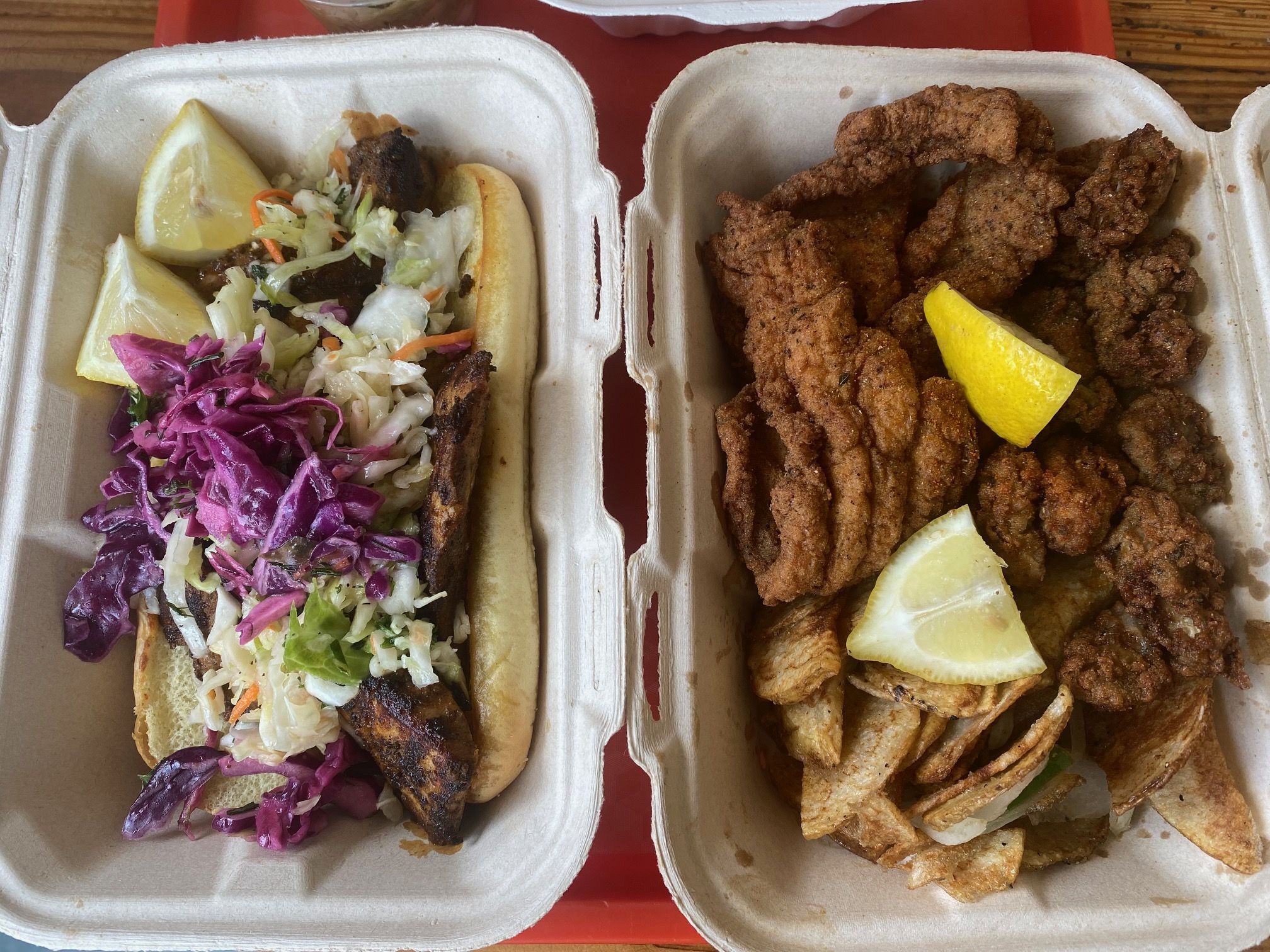 Two compostable containers on a wooden surface with fried fish and potato wedges with lemon wedges in one, and a fish sandwich topped with coleslaw and lemon wedges in the other.