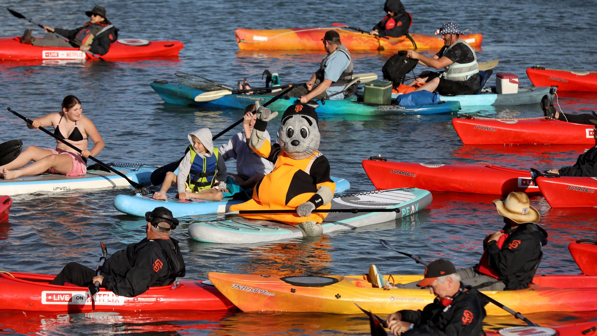 Several people paddle kayaks and stand-up paddleboards on a calm lake. In the center, a person in an orange life jacket sits on a board with a panda mascot head, surrounded by colorful kayaks.