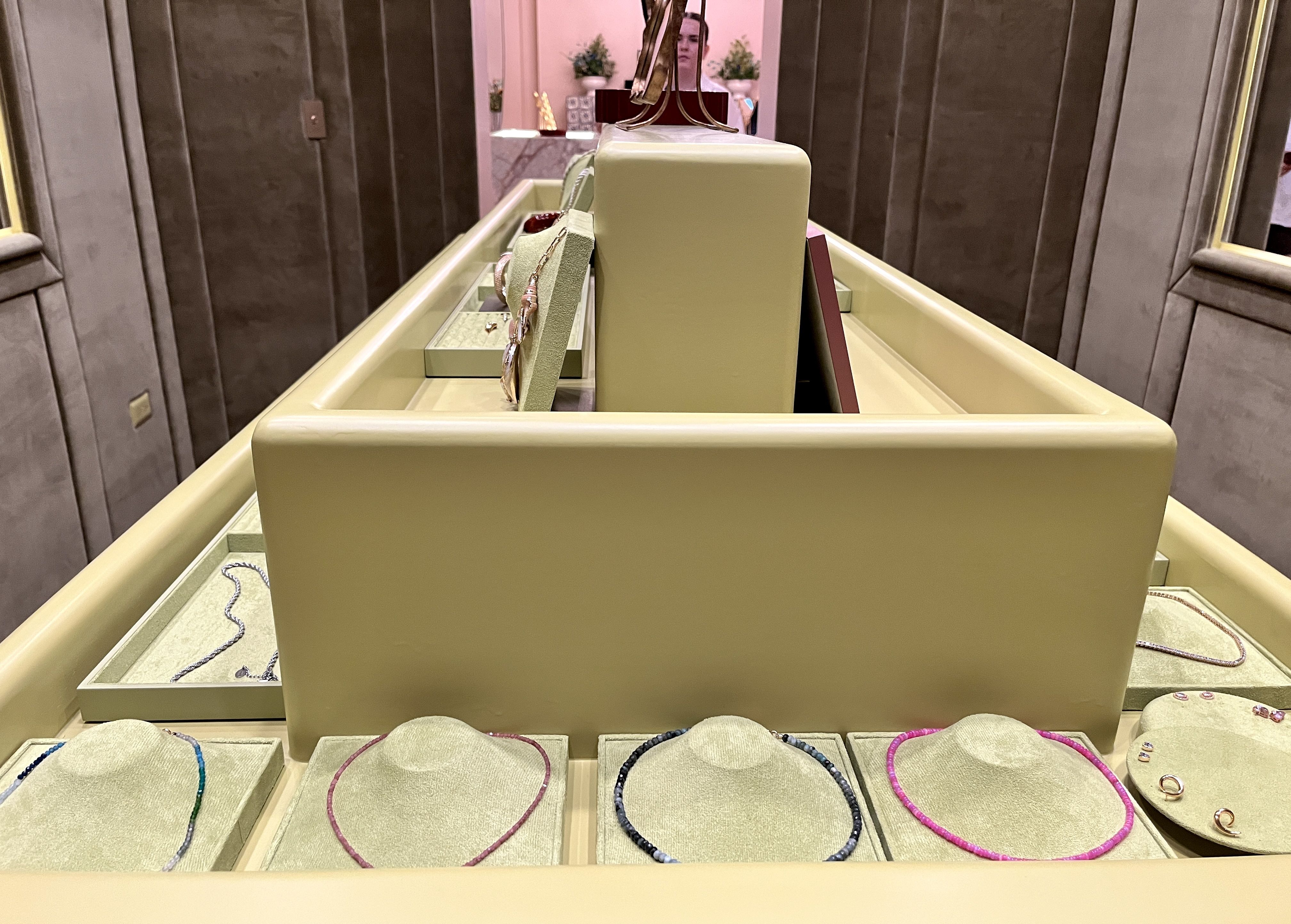 Wide shot of a jewelry display: pale yellow-green cases hold bracelets and rings on padded blocks; a central pedestal supports a decorative metal piece, with a shopper visible in a pink background.