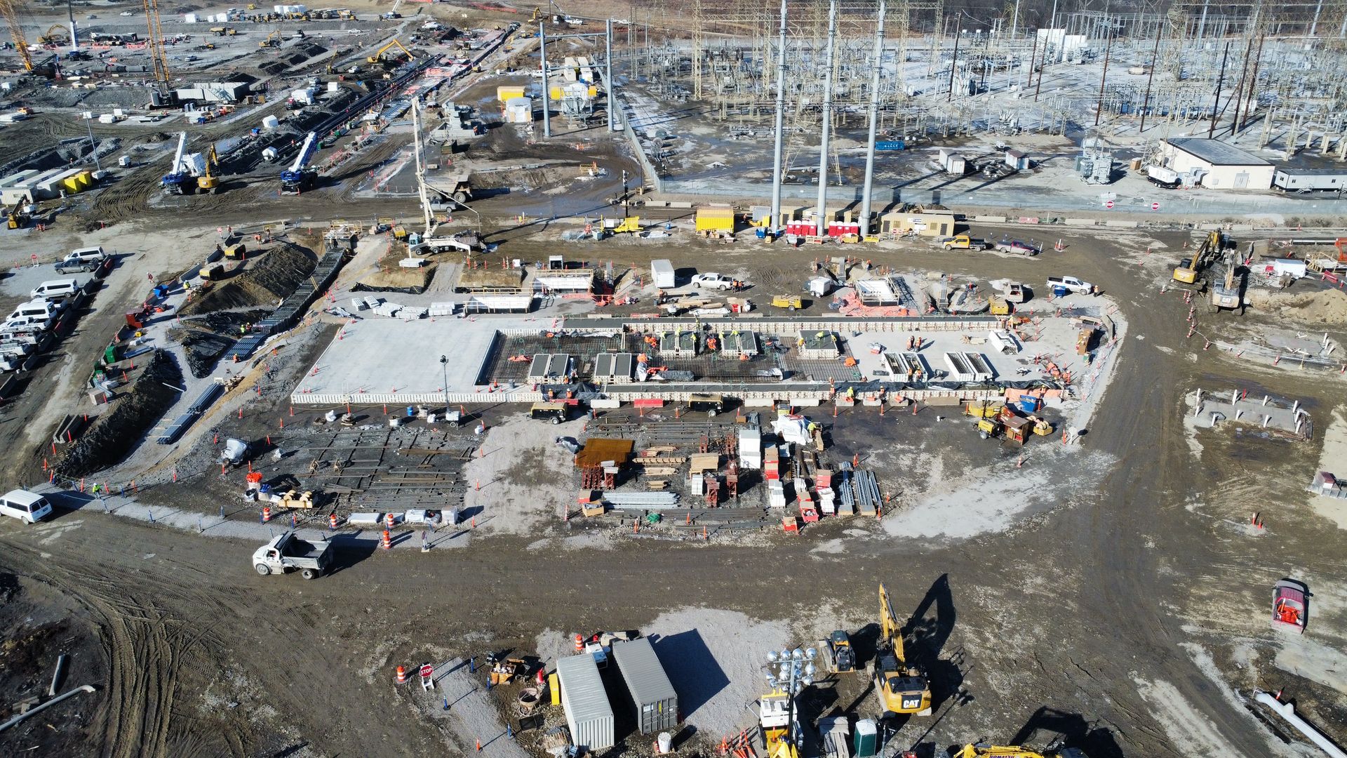 Aerial view of a large construction site with cranes, heavy machinery, and workers. Concrete foundations, steel rebar grids, and piles of lumber surround a central building area; dusty, muddy ground.