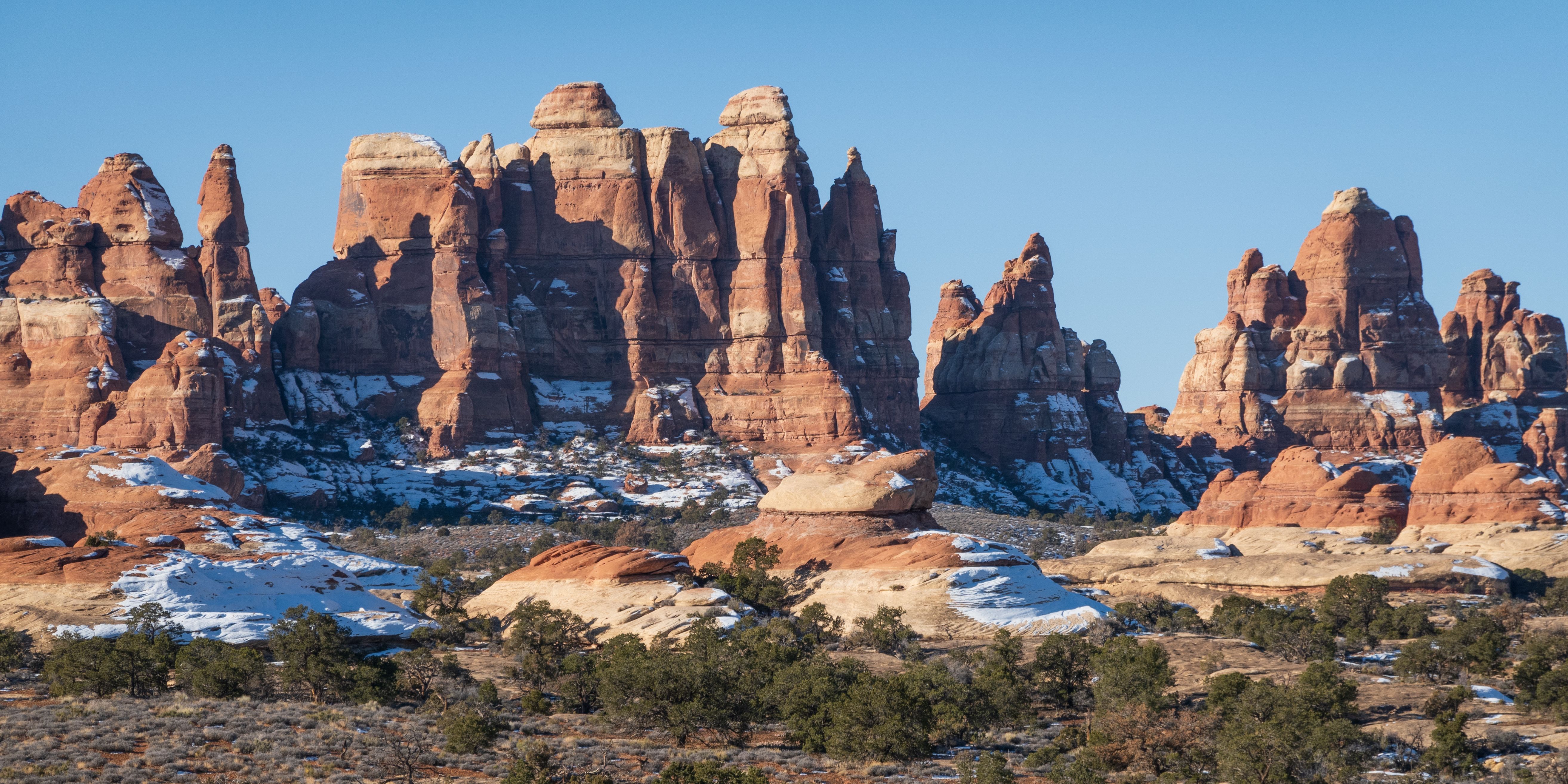 Red rock formations with patches of snow under a clear blue sky, surrounded by sparse green trees and dry vegetation in a desert landscape.
