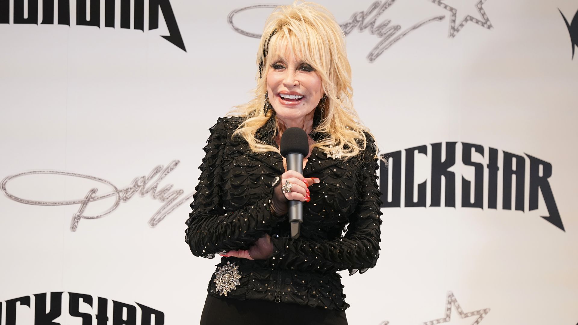 Dolly Parton, smiling, holding a microphone, wearing a shiny black jacket