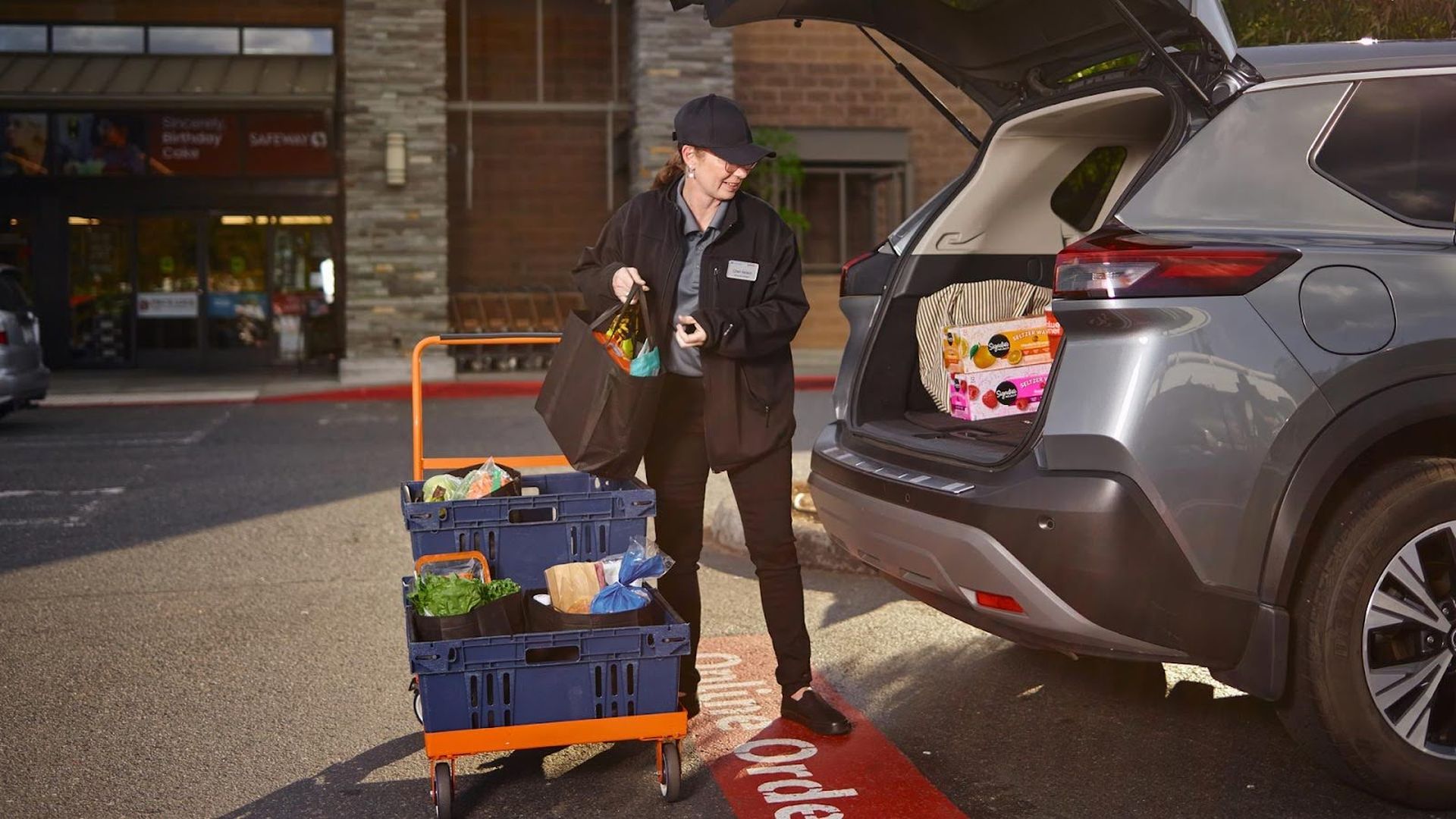 A person wearing a black cap and jacket loading grocery bags from a blue cart into the open trunk of a silver SUV in a parking lot outside a store.