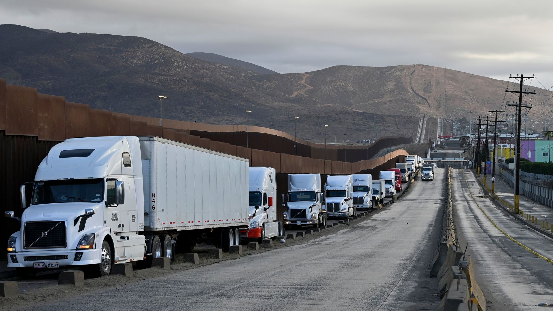 Line of white and red semi trucks parked next to a tall, brown border wall with mountains in the background under a cloudy sky.