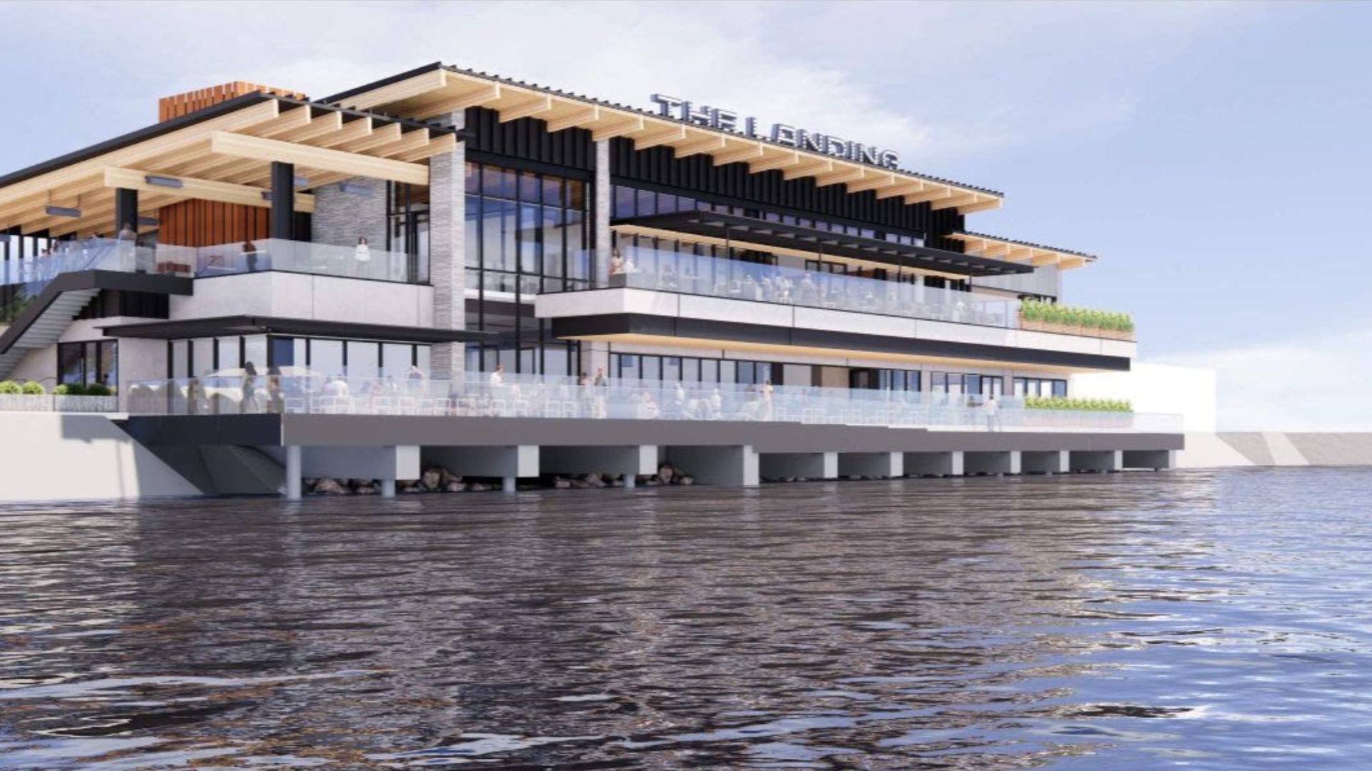 Modern multi-level waterfront building named "The Landing" with large glass windows, wooden beams, balconies with glass railings, and people visible inside and outside near calm water.