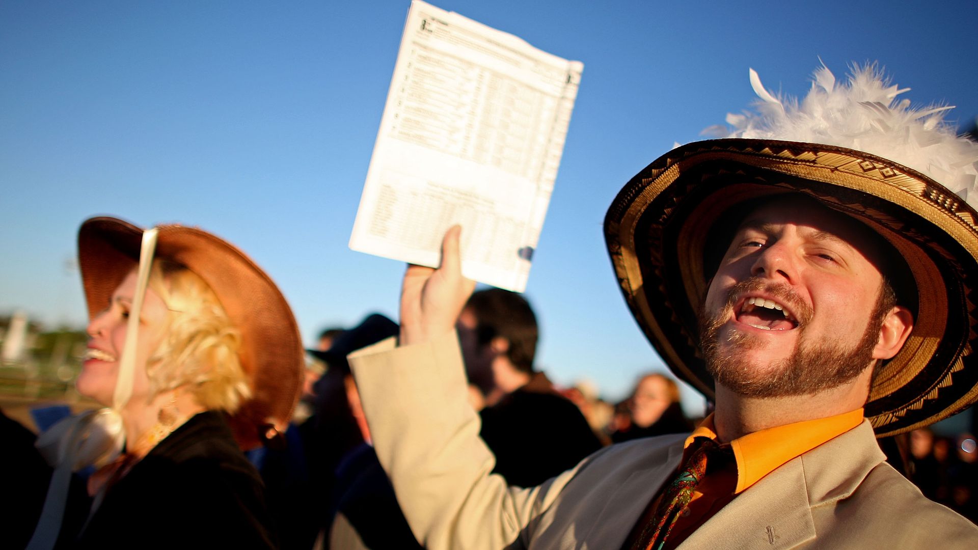 Man wearing a beige suit, orange shirt, patterned tie and feathered hat enthusiastically holding up a racing program. 