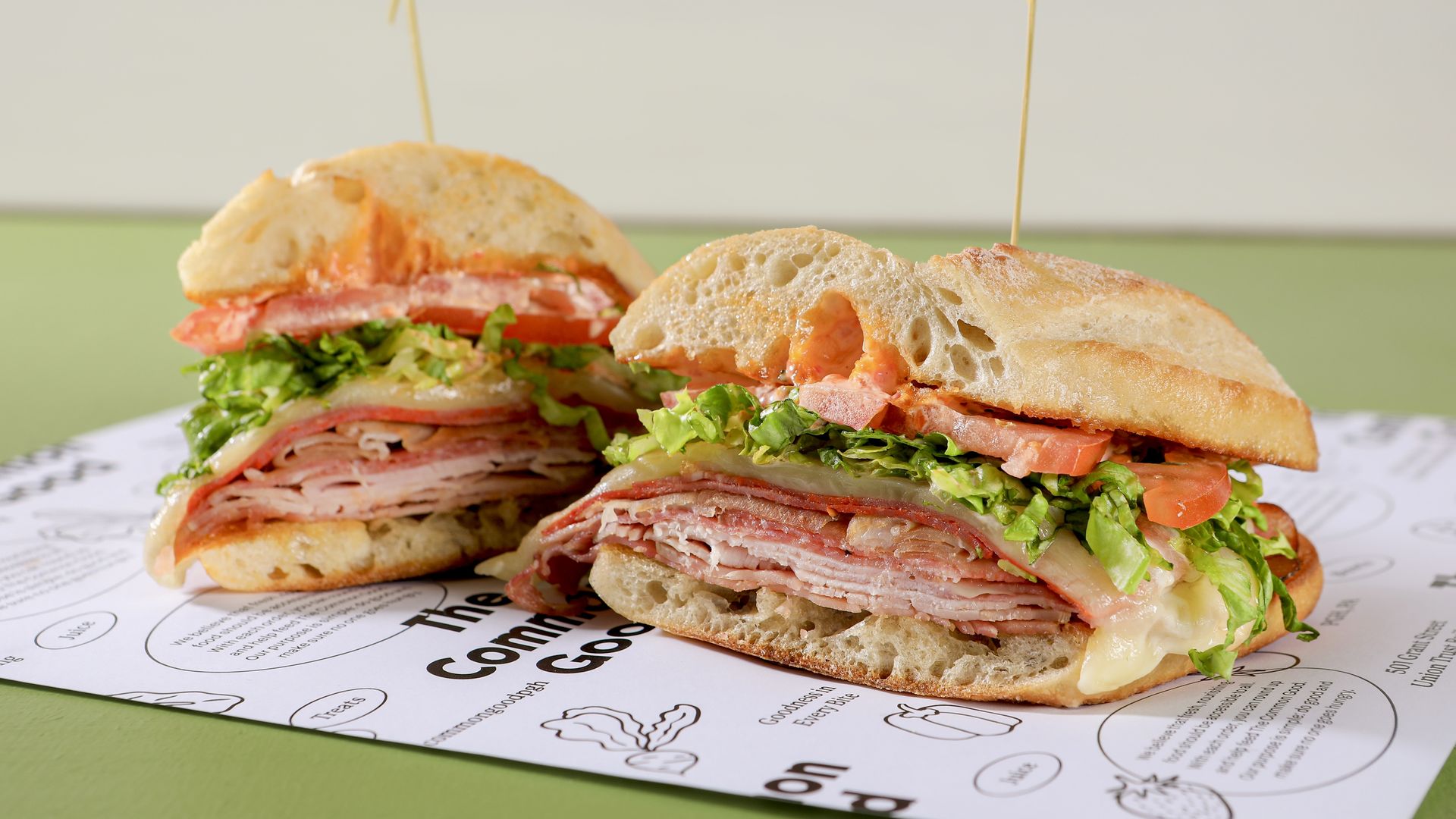 Two halves of a sandwich with layers of sliced deli meats, melted cheese, lettuce, and tomato on airy ciabatta bread, held with wooden picks on a printed paper background.