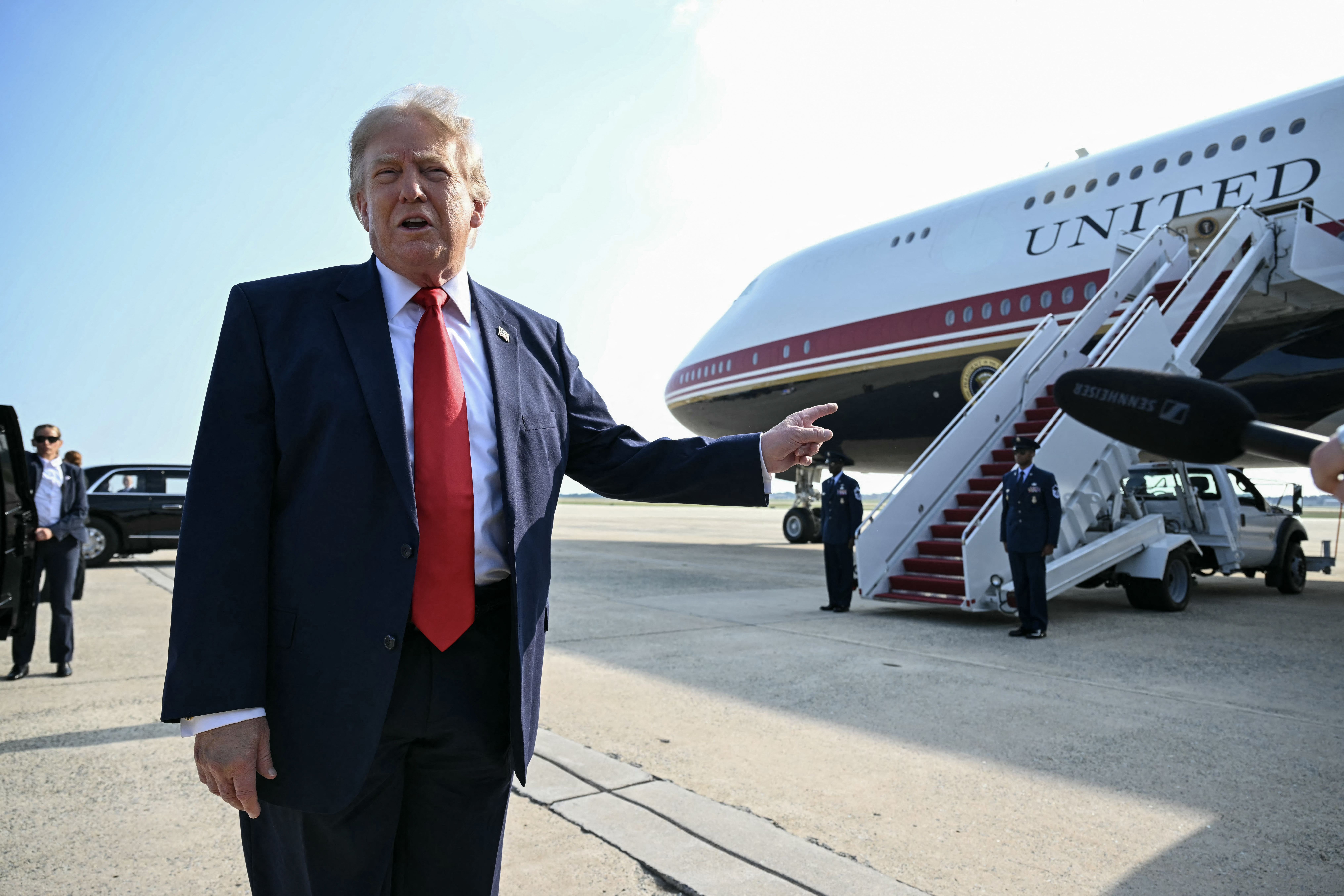 President Trump — wearing a dark suit, a white collared shirt, a red tie and an American flag pin — points at Air Force One as he speaks to members of the press.