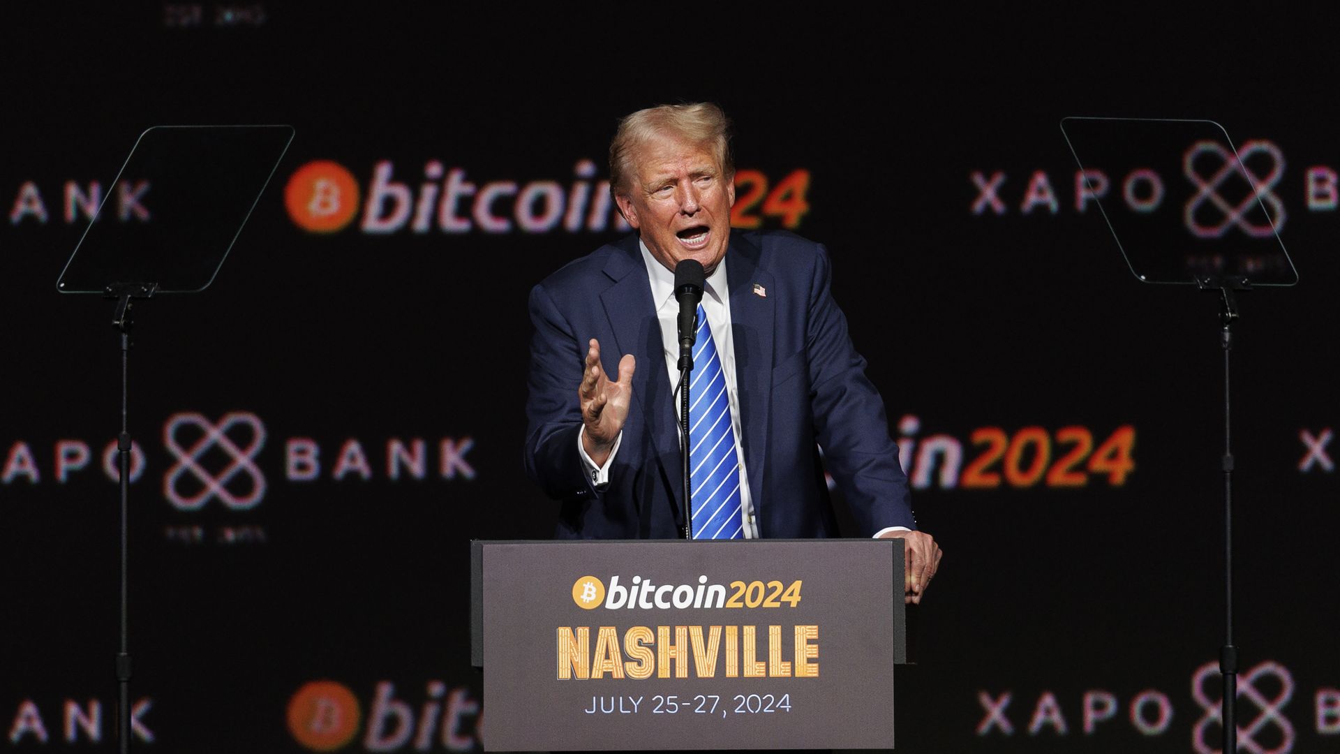 Man in a dark suit and striped tie speaks into a microphone at a podium on a dark stage. Behind him are Bitcoin 2024 Nashville banners and logos; the podium reads Bitcoin 2024 Nashville July 25-27, 2024.