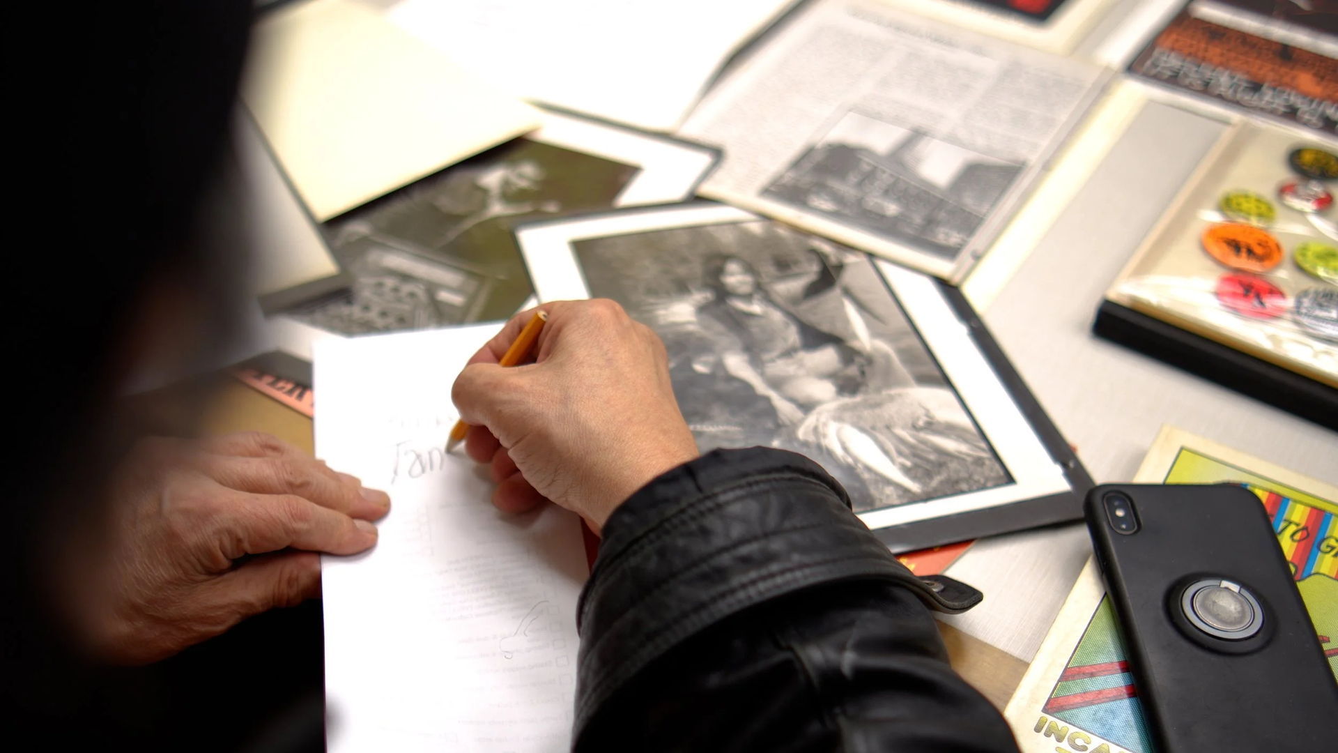 Close-up of a person in a black jacket writing on a white sheet with a pencil at a cluttered desk. Scattered photos, magazines, and a smartphone surround the pages.