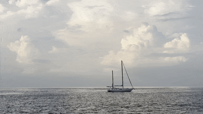 A lone sailboat with two masts sails on calm dark blue-gray water under a vast sky filled with large, white, fluffy clouds.
