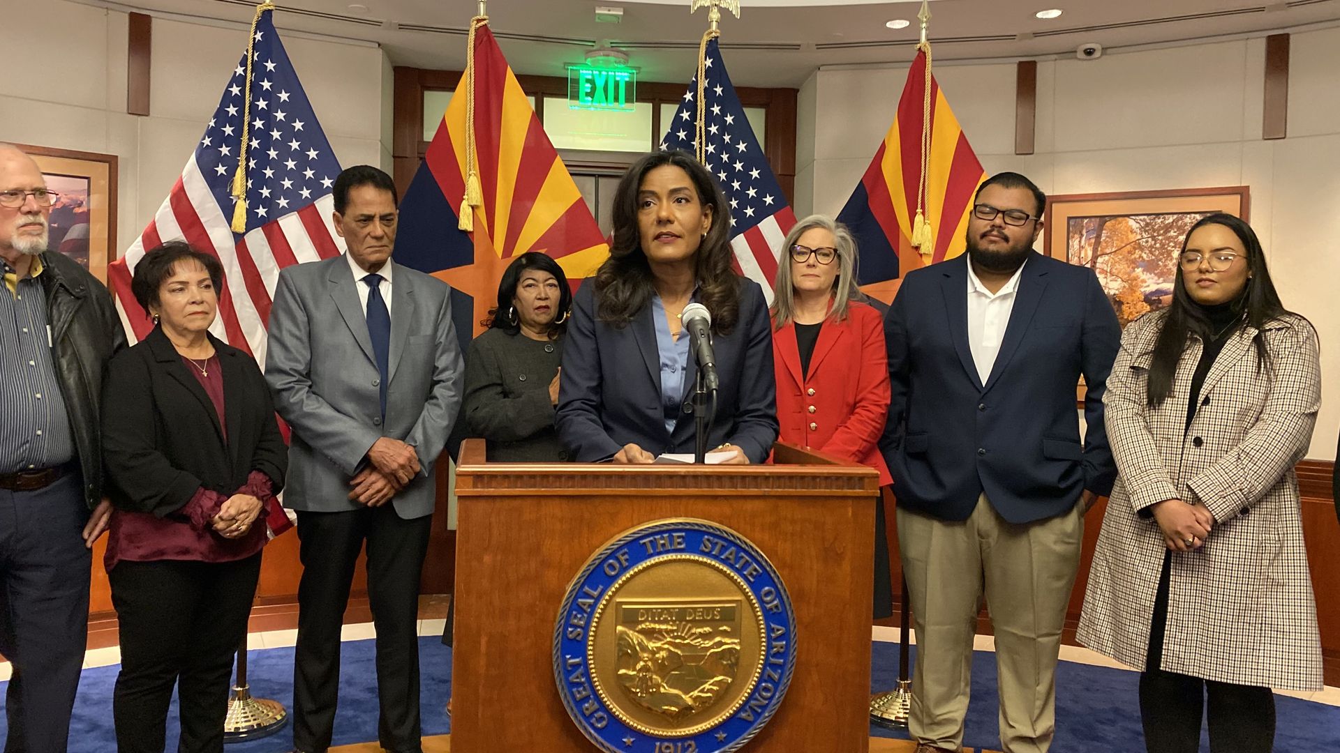 Maria Elena Cruz speaks at a lectern while members of her family and Governor Katie Hobbs stand behind her. 