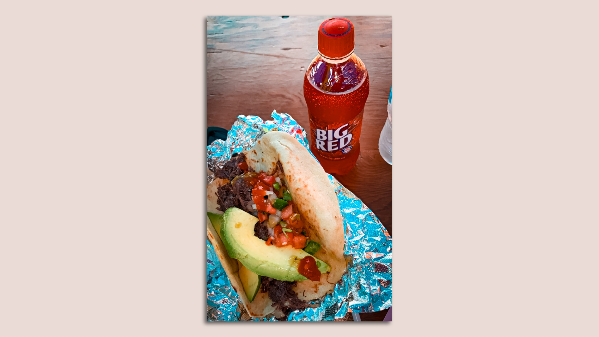A tortilla filled with barbacoa meat that's topped with chopped onions, tomatoes and avocado wedges next to a bottle of red soda. 