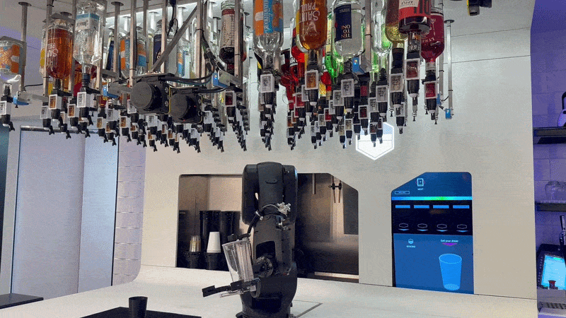 Bar scene with numerous liquor bottles hanging from the ceiling, a black beverage dispenser on a white counter, and a blue digital drink menu on the wall in the background.