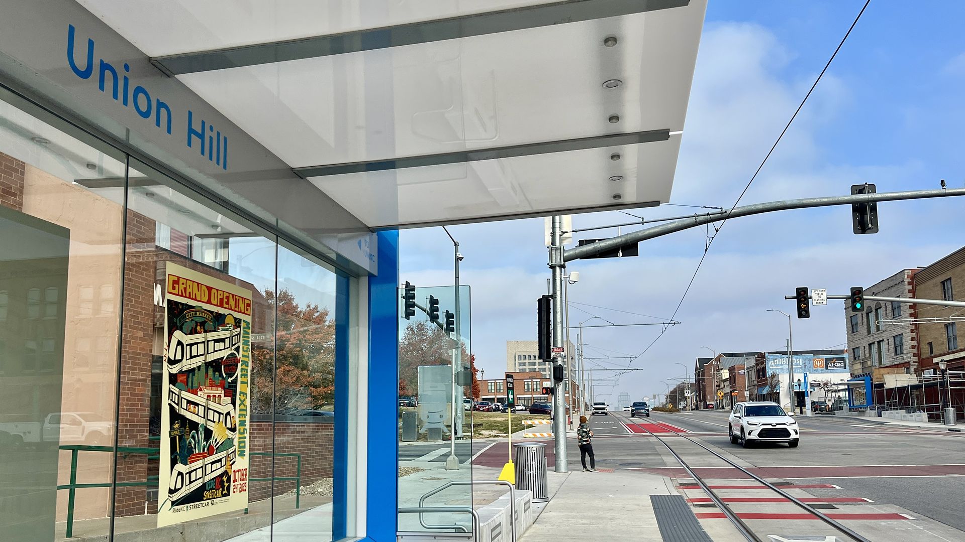 Union Hill streetcar station with glass shelter and a colorful poster announcing a streetcar extension grand opening, street with cars, traffic lights, and a person waiting at the curb.