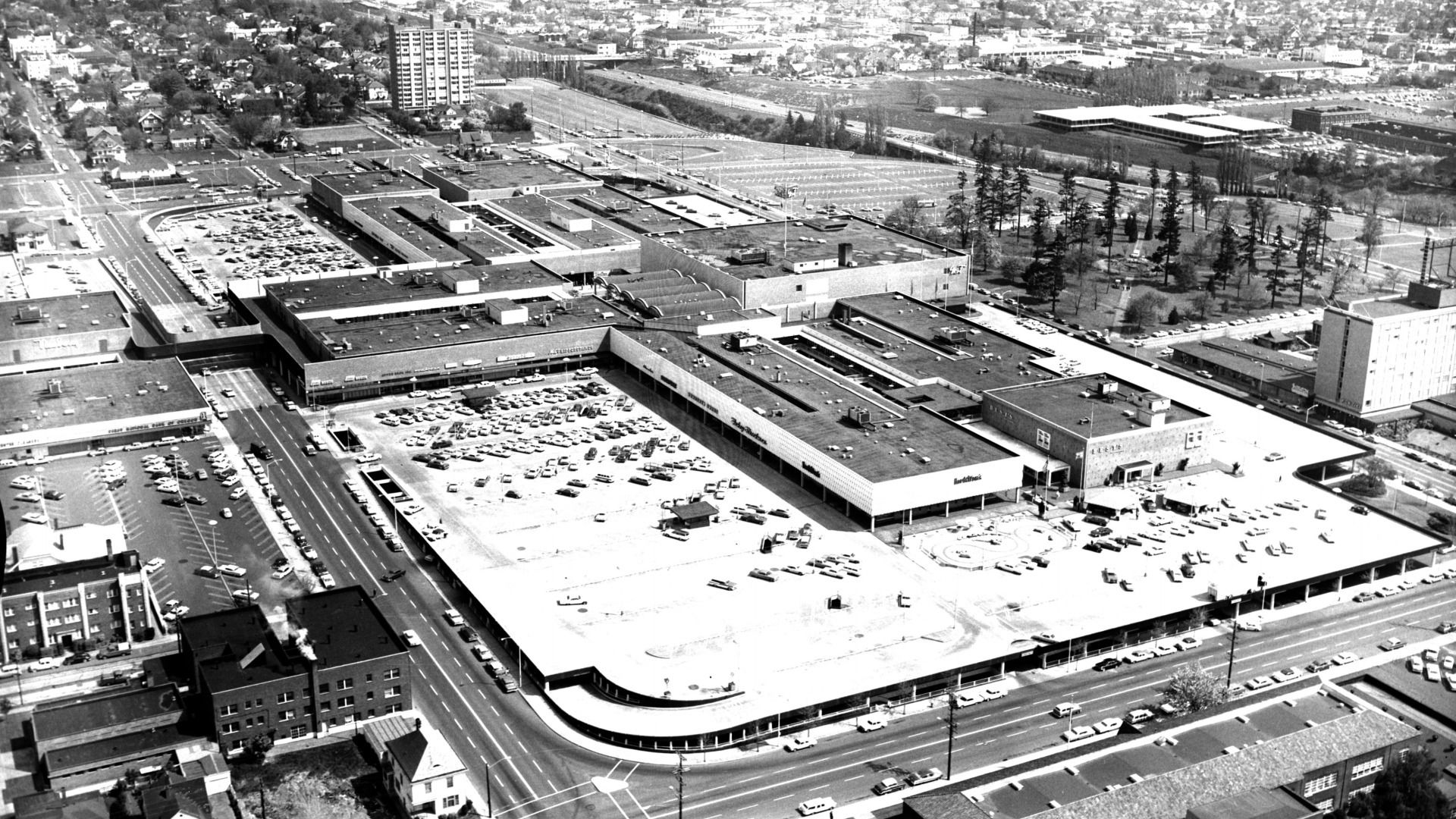 Black-and-white aerial photo of a large shopping mall with extensive parking lots, surrounding city streets, residential area, tall buildings, and a park with trees nearby.