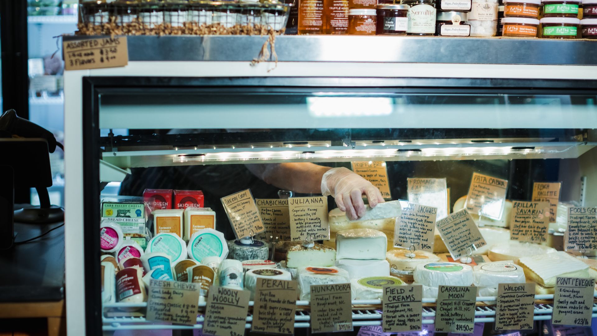 A hand wearing a glove selects a cheese from a glass display case filled with various cheeses, each labeled with handwritten signs describing names and details. Jars of jam are on shelves above.