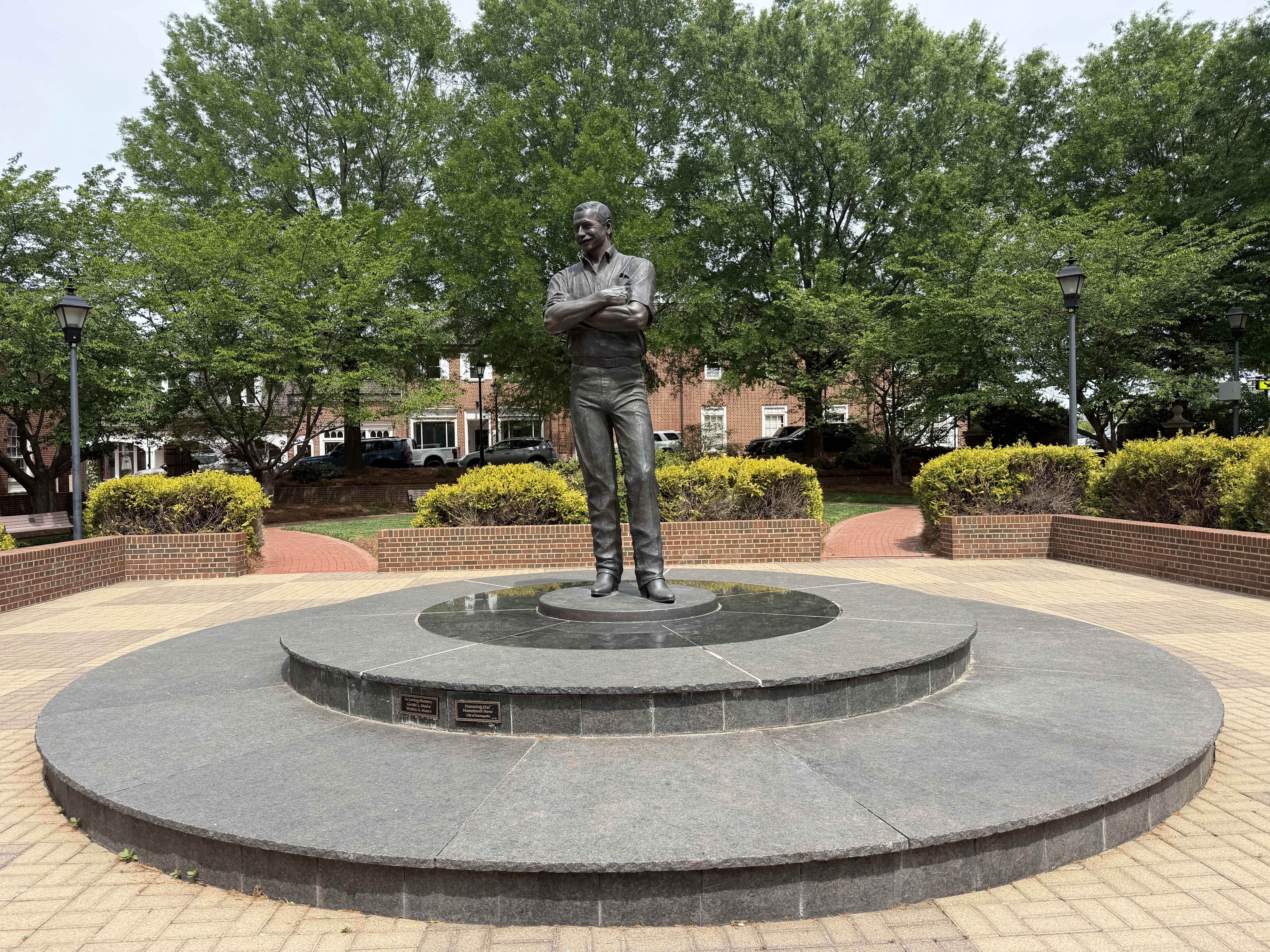 Bronze statue of a man with arms crossed, standing on a circular stone pedestal in a brick-paved plaza. Lush green trees, trimmed shrubs, and street lamps surround the monument.