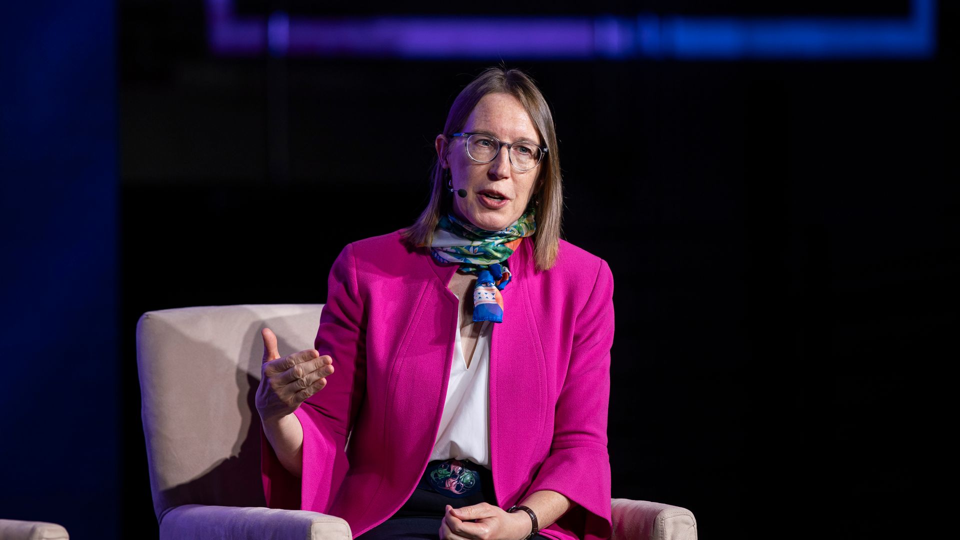 A woman sitting and speaking at an official event, with mid-length hair, glasses, a multi-colored scarf and a bright pink jacket. 