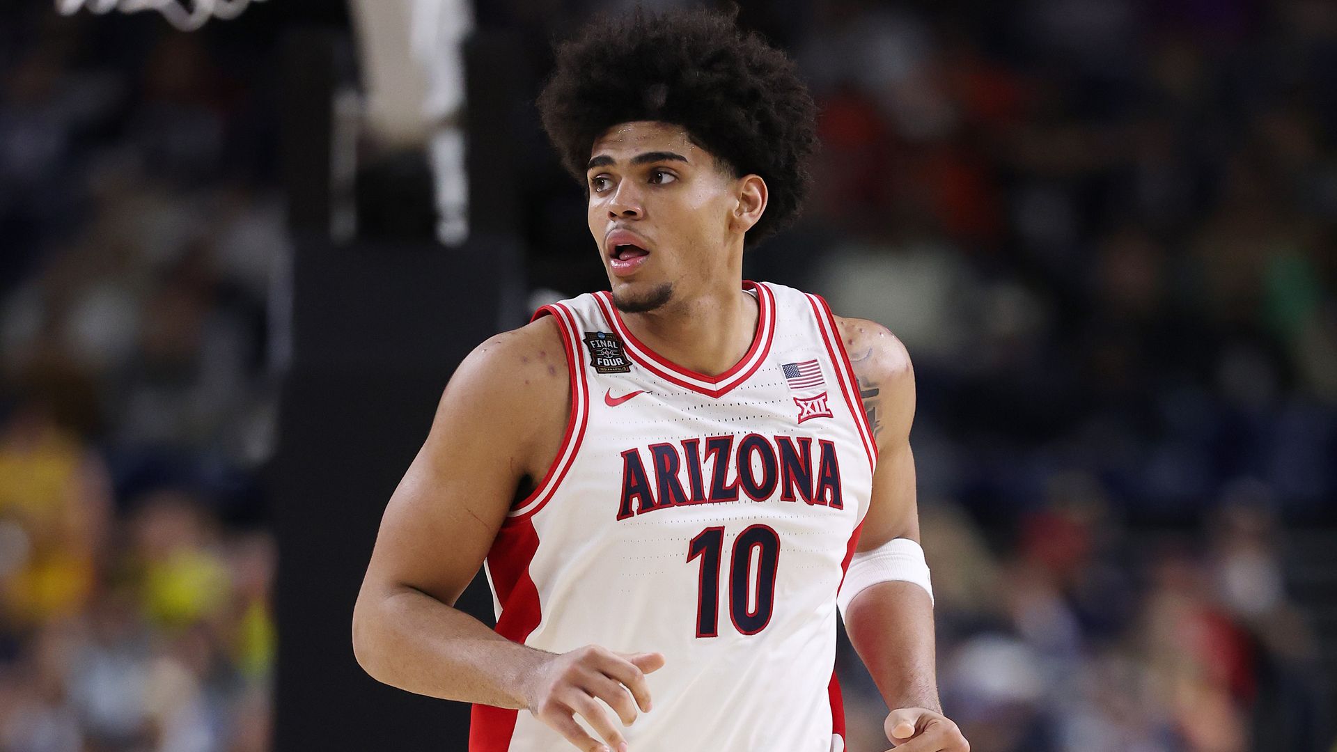 A basketball player in a white uniform with red trimming, with "ARIZONA" and the number 10 on the front of his jersey, jogs up the court while looking to his right.
