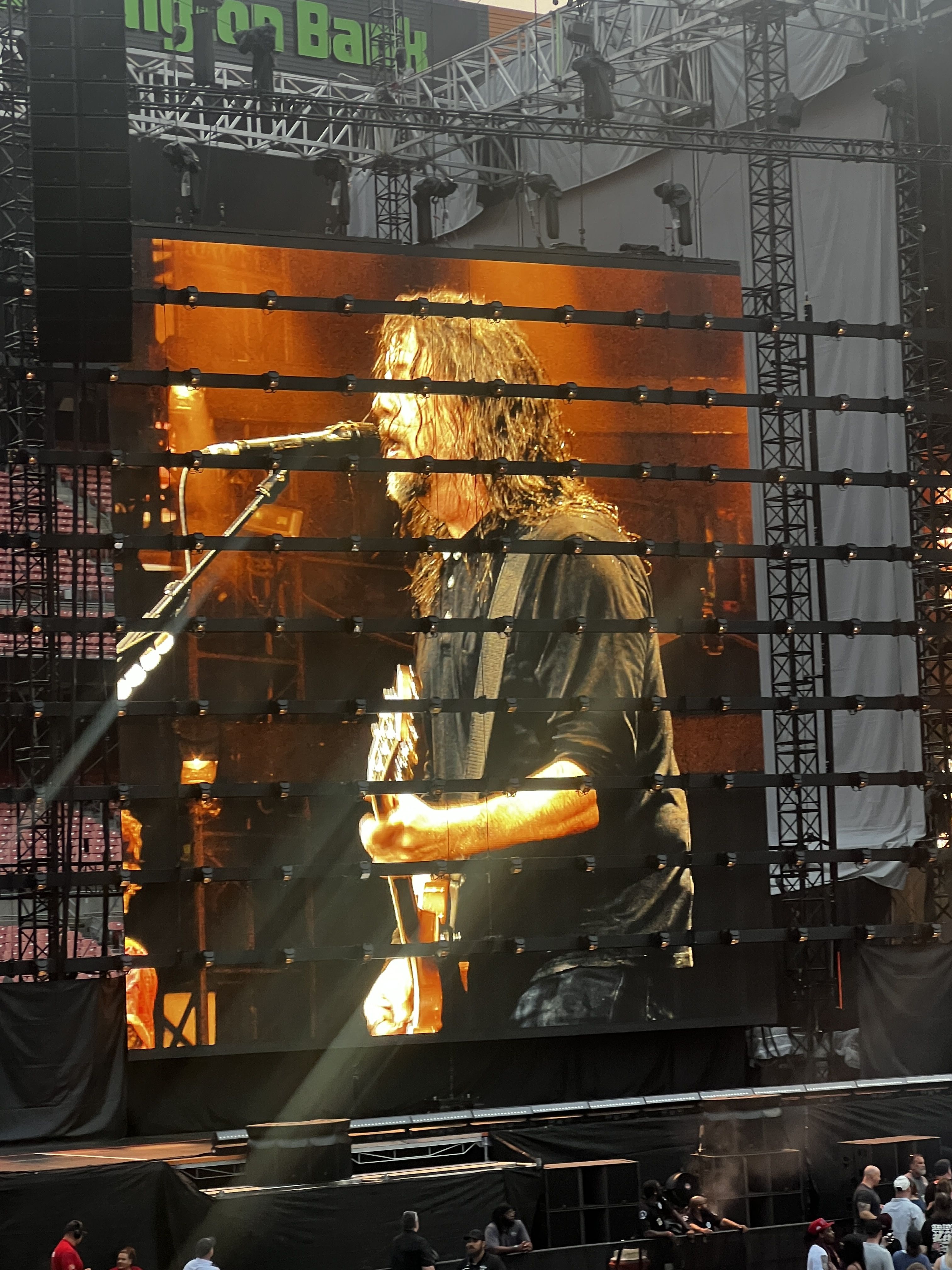 Concert stage with a large orange video screen showing a guitarist with long hair playing a guitar; black scaffolding, stage lights, and a few audience members in front.