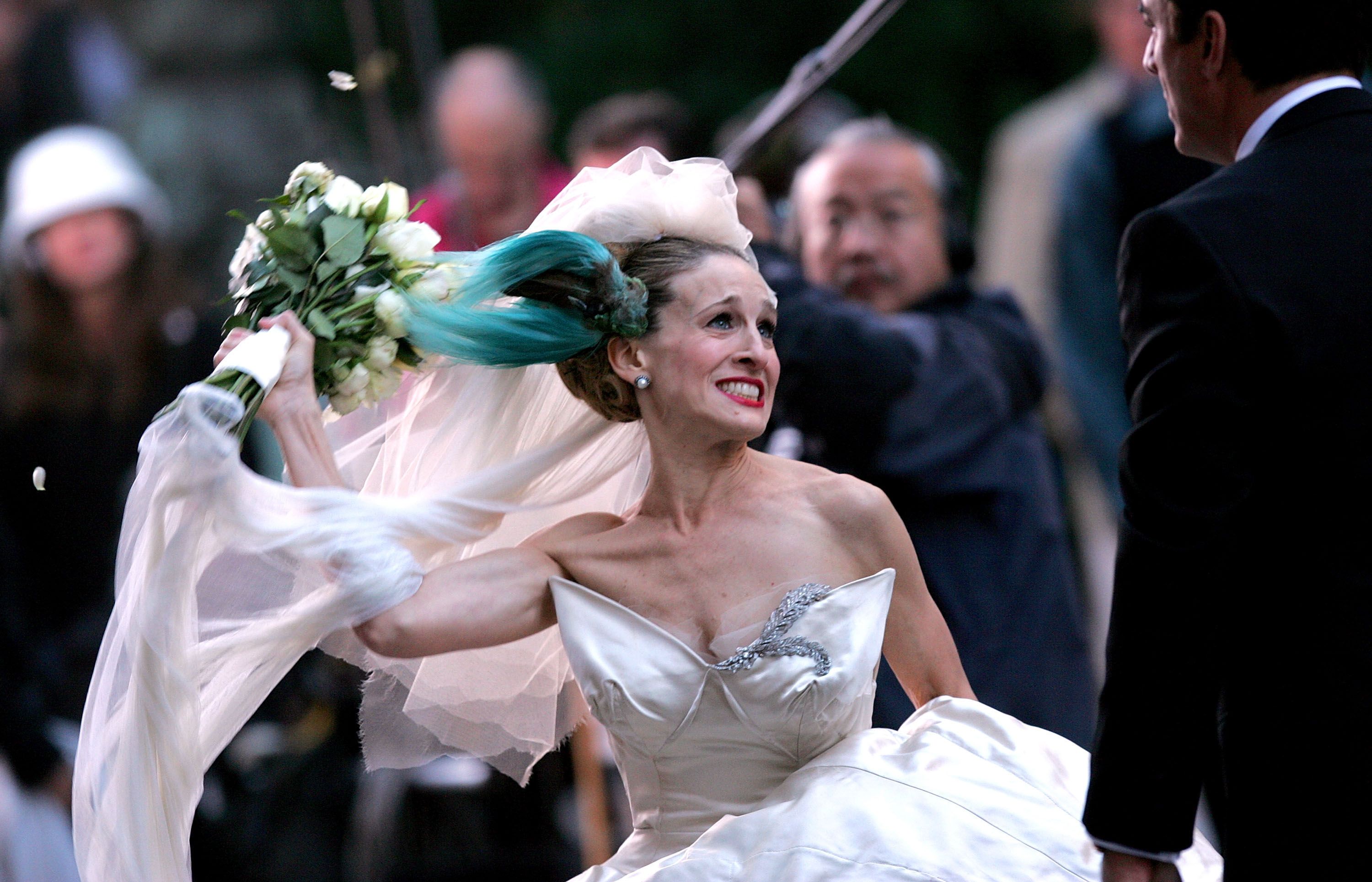 Sarah Jessica Parker, as Carrie Bradshaw, in a white wedding dress angrily swings a bouquet of white roses, facing a man in a black suit
