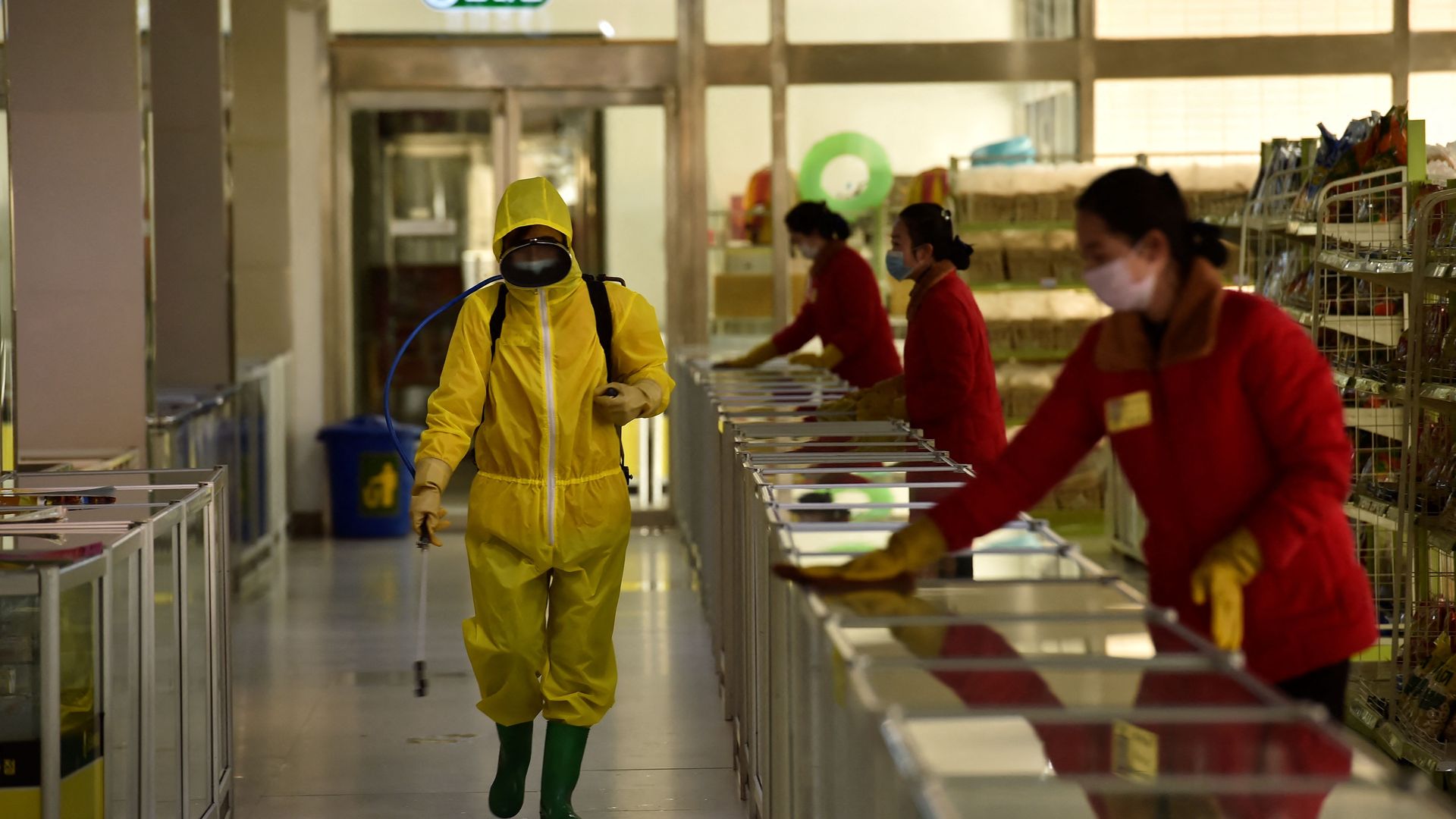 Photo of a person in a yellow hazmat suit walking by a row of counters where people are wiping down surfaces