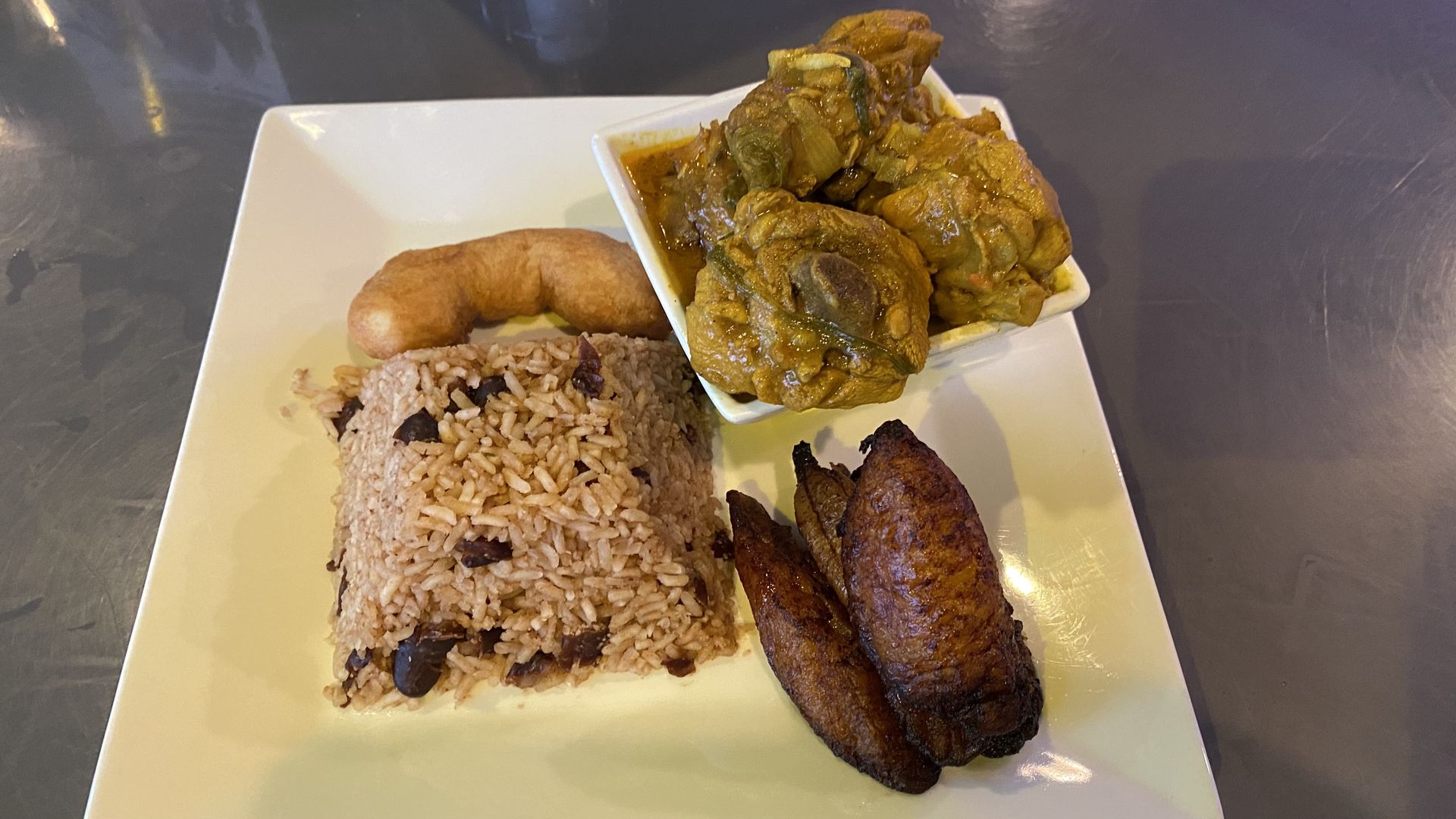A square plate with rice and beans, fried plantains, round fried bread and a bowl of curry chicken.