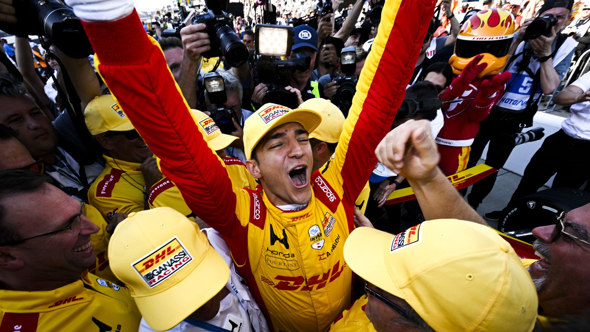 Celebration scene with photographers crowded around a jubilant race driver in a yellow DHL racing suit, arms raised in victory; yellow caps, bright gear, and a flame-painted helmet among the cheering crowd.