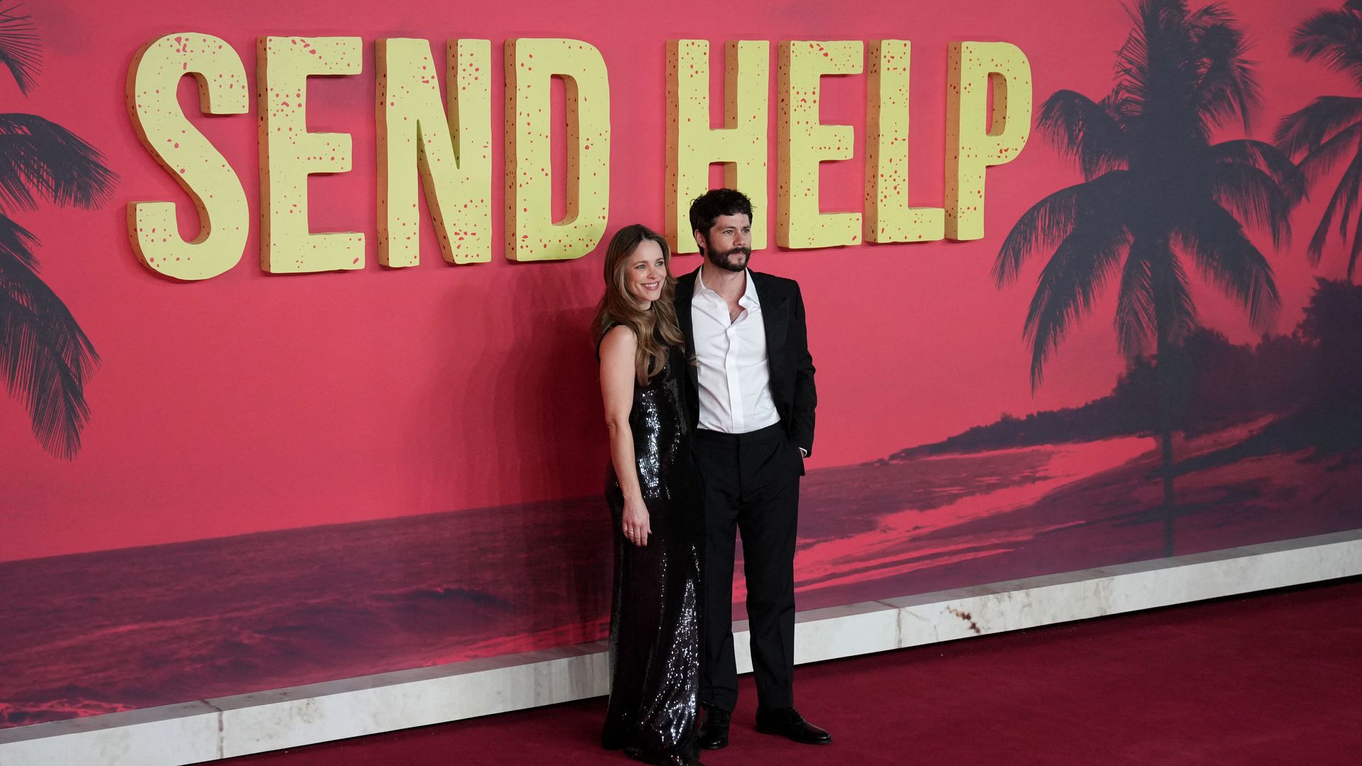 Rachel McAdams in a sparkly black gown and Dylan O'Brien in a black suit with white shirt stand together in front of a "SEND HELP" sign.