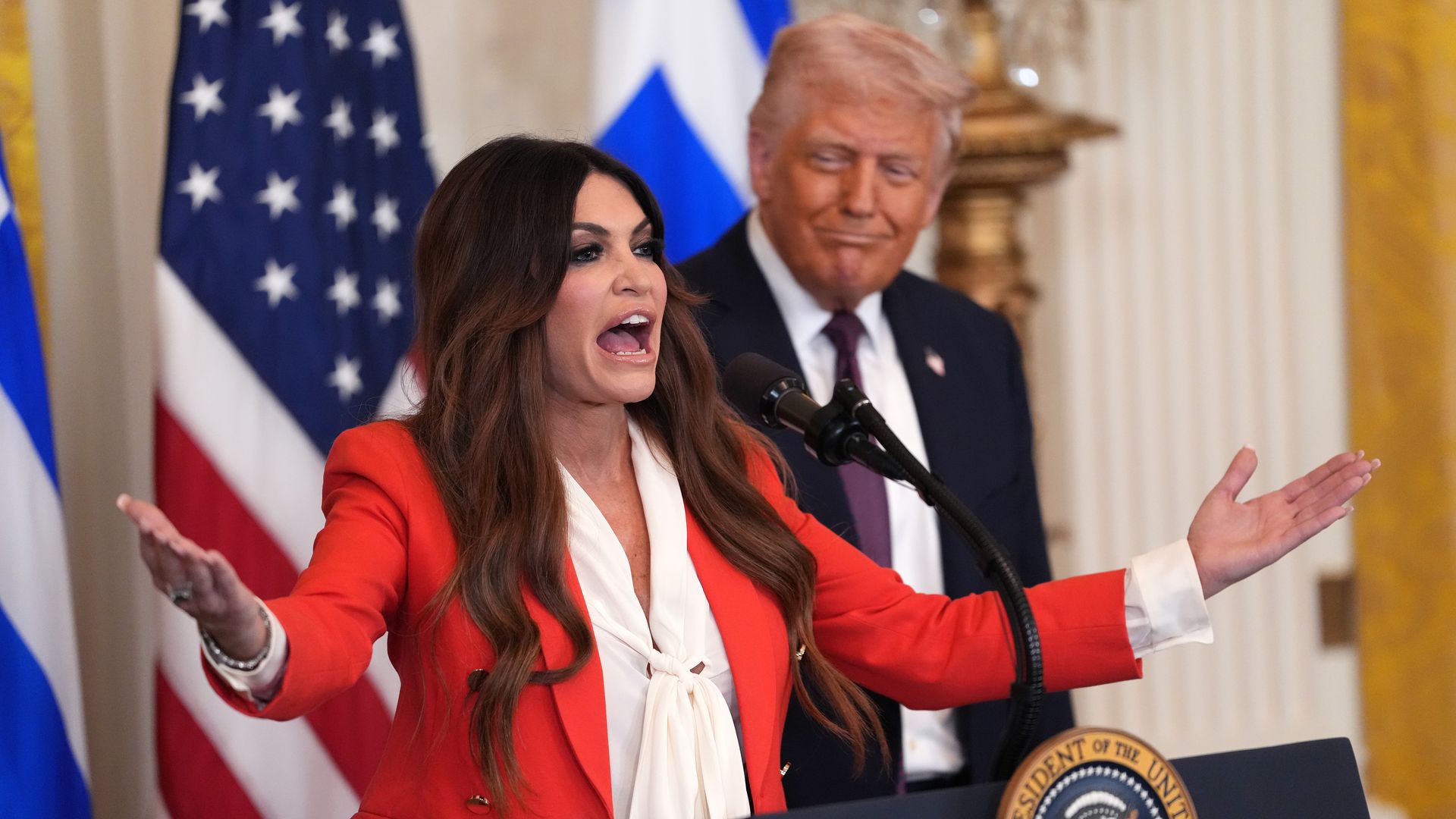 A long, dark-haired Kimberly Guilfoyle, the newly confirmed U.S. ambassador to Greece, wearing a bright red coat and white blouse, gestures as she speaks into a mic at a podium as a smiling President Trump, wearing a navy jacket with a US flag pin, a white shirt and purple tie, looks on.