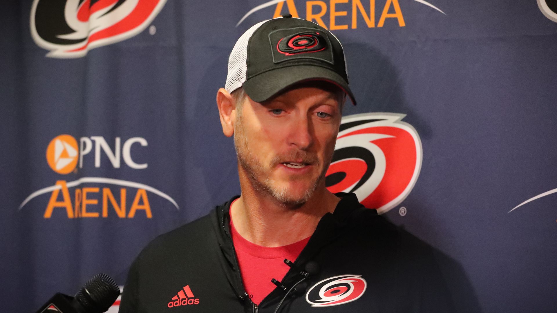 Man with light beard wearing a black and white Carolina Hurricanes cap and black Adidas jacket speaking in front of a backdrop with Carolina Hurricanes and PNC Arena logos.