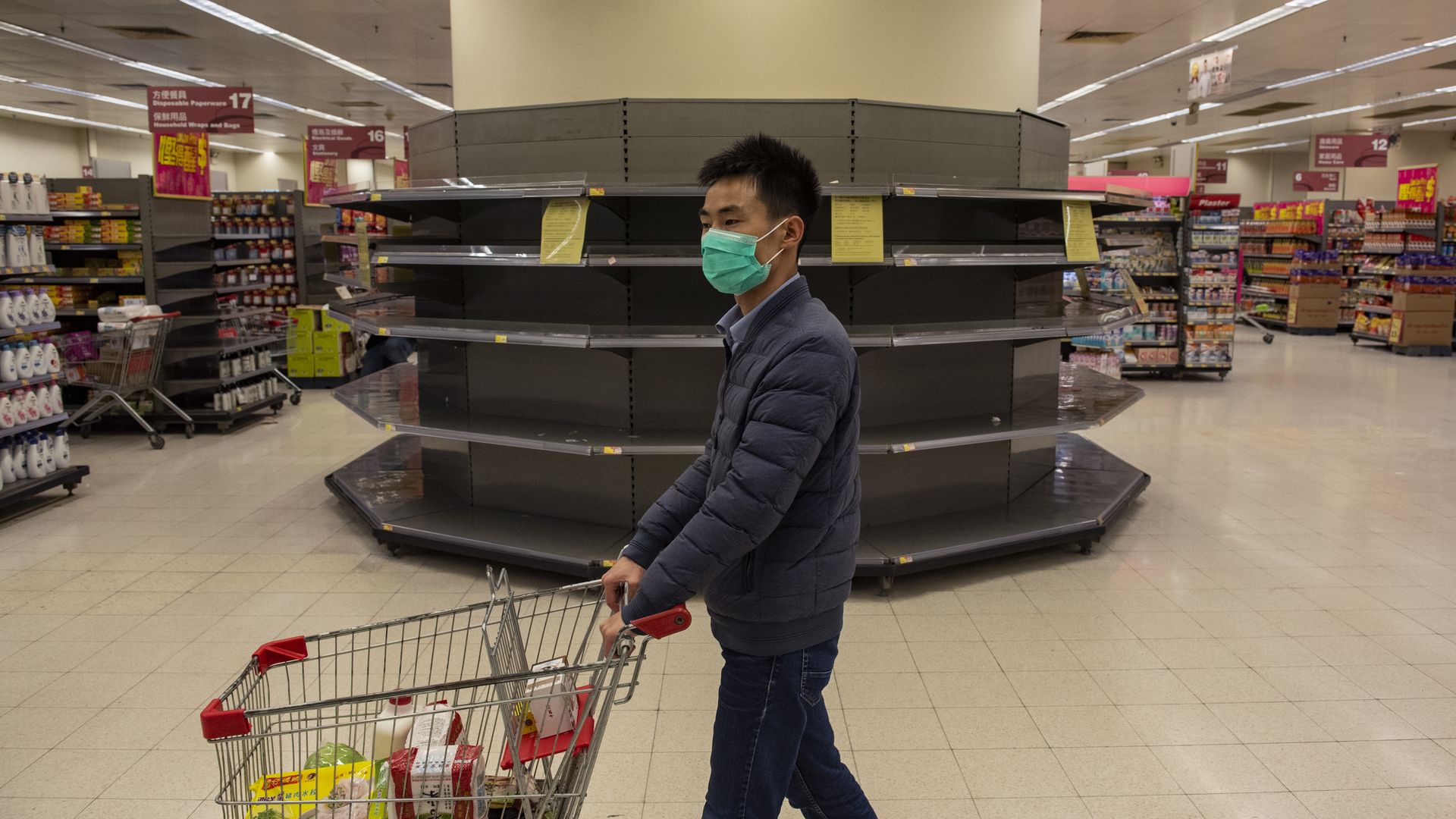 In this image, a man pushes a shopping cart past empty shelves while wearing a face mask.