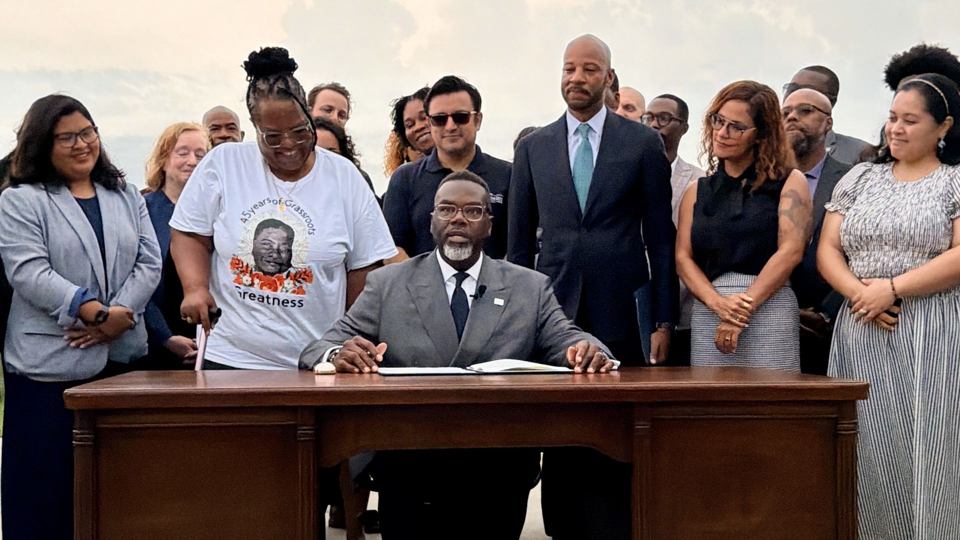 Group of diverse people surrounding a large wooden desk as a man in a gray suit signs papers; others in business attire stand outdoors, under a pale cloudy sky.