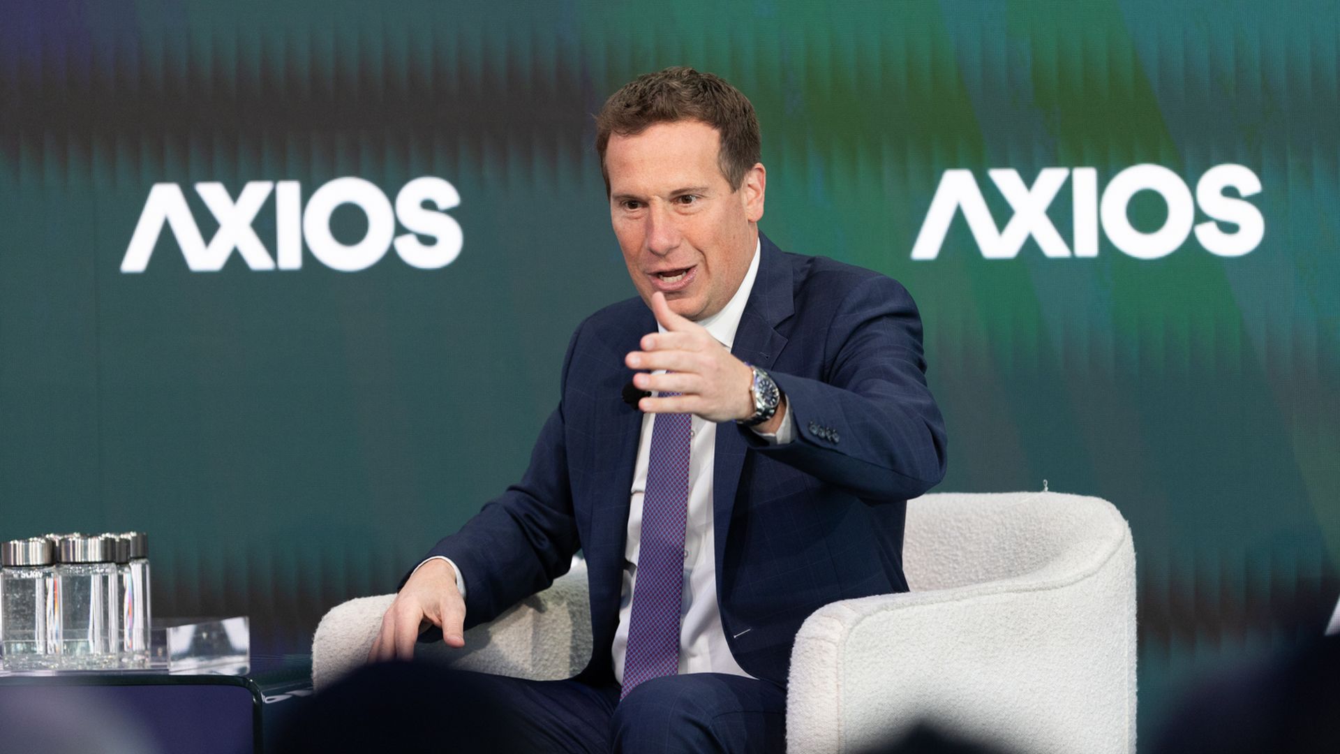 Man in a navy suit and purple tie sits in a white chair on stage, gesturing with his right hand while speaking. Behind him, a green Axios backdrop shows the word AXIOS.
