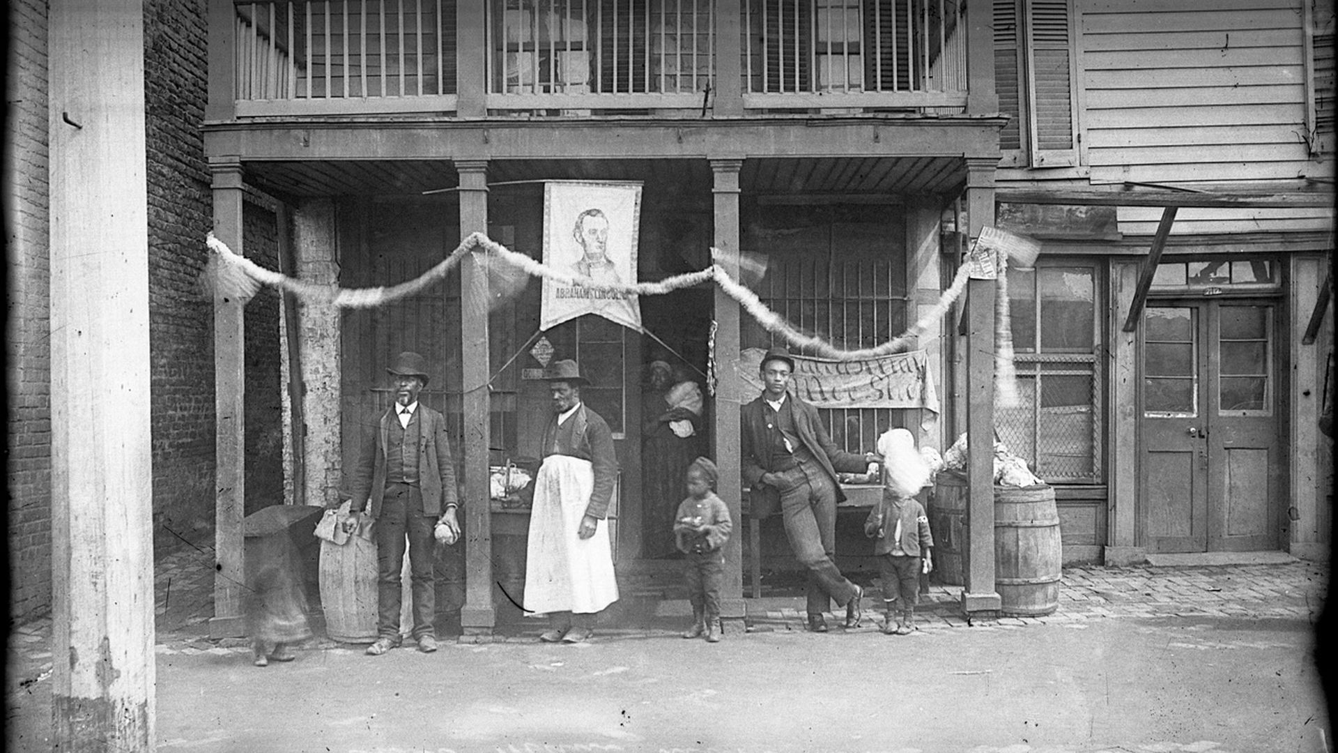some people in the 1800s - 2 men, 2 women, 2 kids and a baby - stand outside what looks like and old timey saloon in Richmond 
