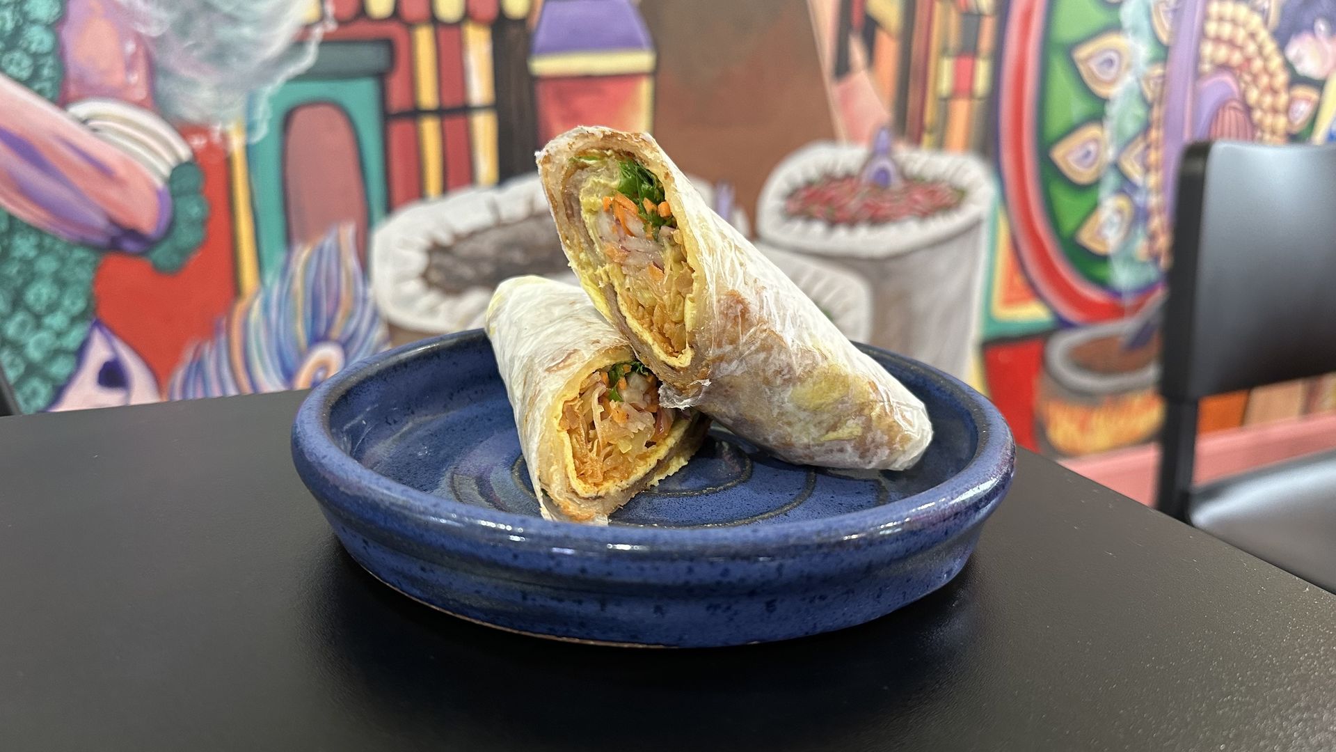An egg kaathi roll sits on a blue plate on a black table in front of a mural.