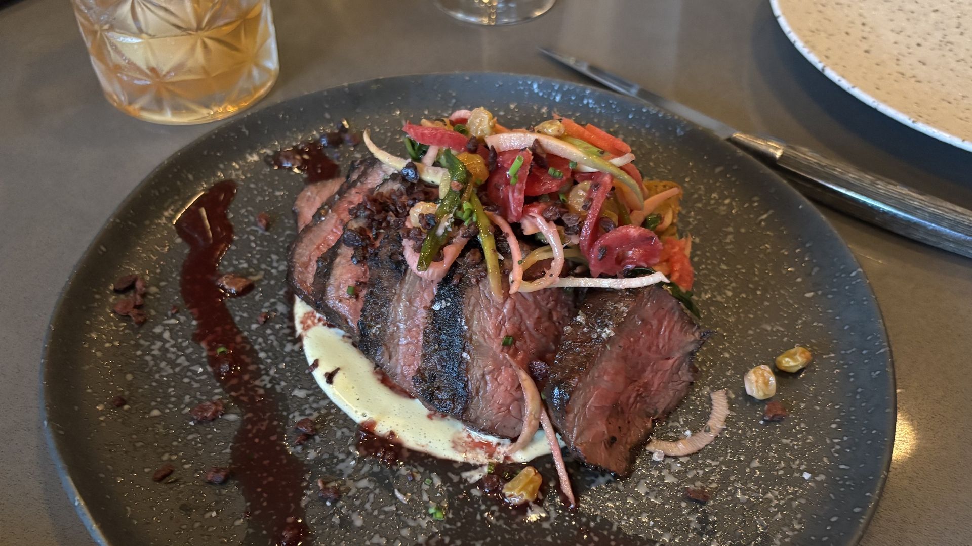 Thinly sliced medium-rare steak topped with a colorful onion, pepper, and herb slaw on a dark speckled plate, with dark sauce, pale sauce smear, and scattered nuts; a glass of amber drink nearby.