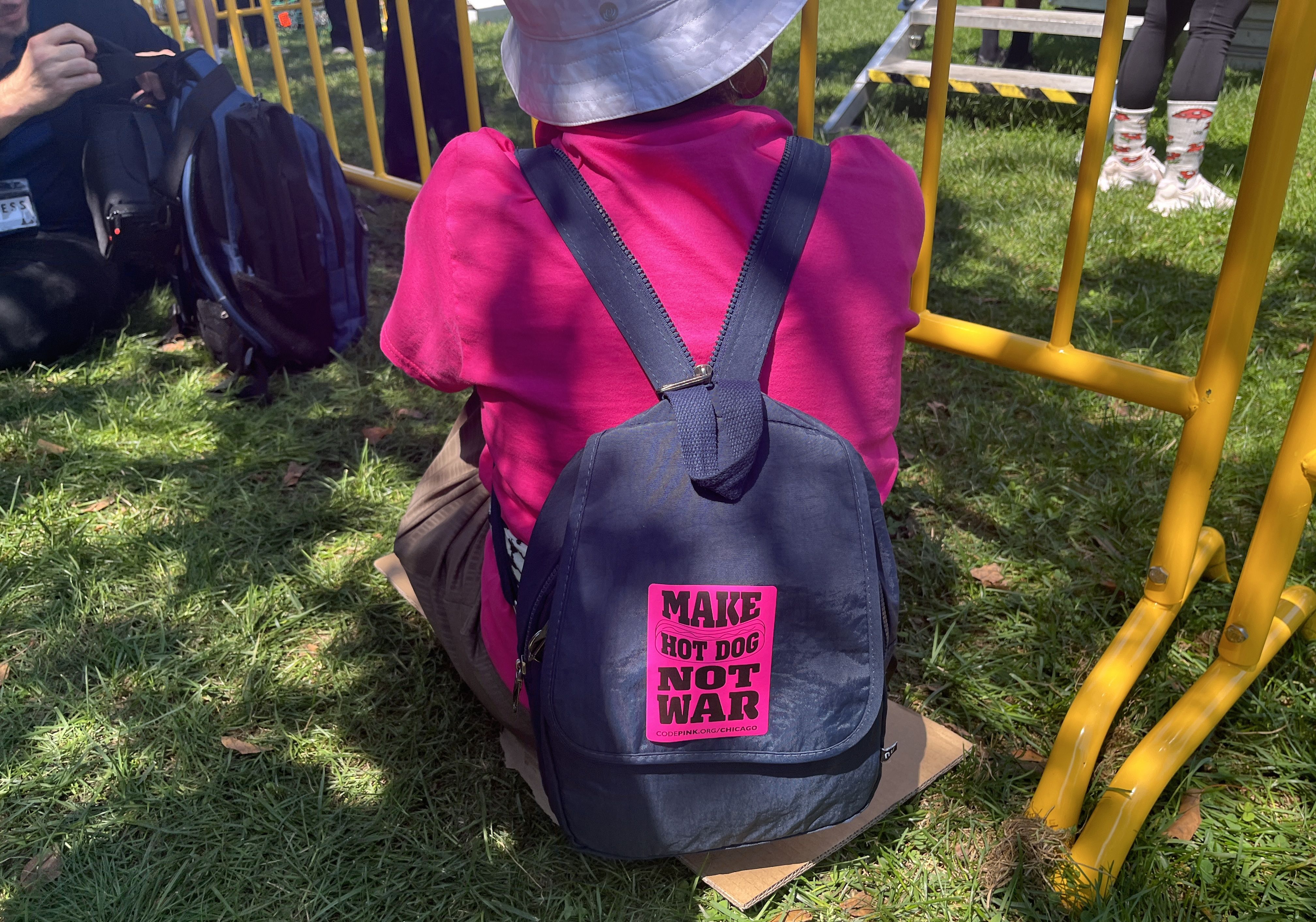 A person wearing a pink t-shirt, wearing a blue backpack with a hot pink sticker that reads "Make hot dog, not war."